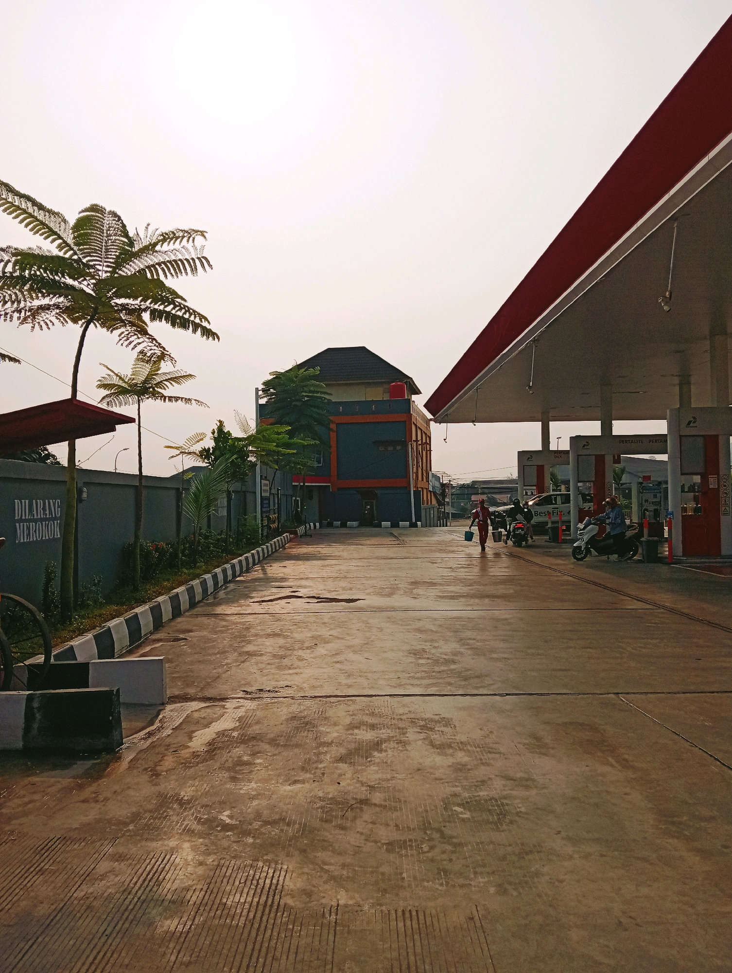 The image depicts a gas station in Samarinda, Indonesia, under a bright, overcast sky. In the foreground, a concrete forecourt is wet with scattered puddles, reflecting the muted light. A low wall borders the left side of the forecourt, with a sign on it reading "DILARANG MEROKOK" (No Smoking) in white text. To the left of the wall, a tall, thin tree with feathery fronds stands, partially obscuring another similar tree behind it.
Further into the scene, under the red-and-white striped canopy of the gas station, several fuel pumps are visible. Two motorcycles are parked near a pump, and one scooter is occupied by a person. A person wearing a red shirt and white pants walks away from the pumps carrying a blue bucket. In the background, a multi-story building with orange and blue accents rises above the gas station structures. Trees and other vegetation are scattered throughout the scene, and power lines are faintly visible in the distance. The overall impression is of a quiet, functional area at a gas station in an urban environment.
