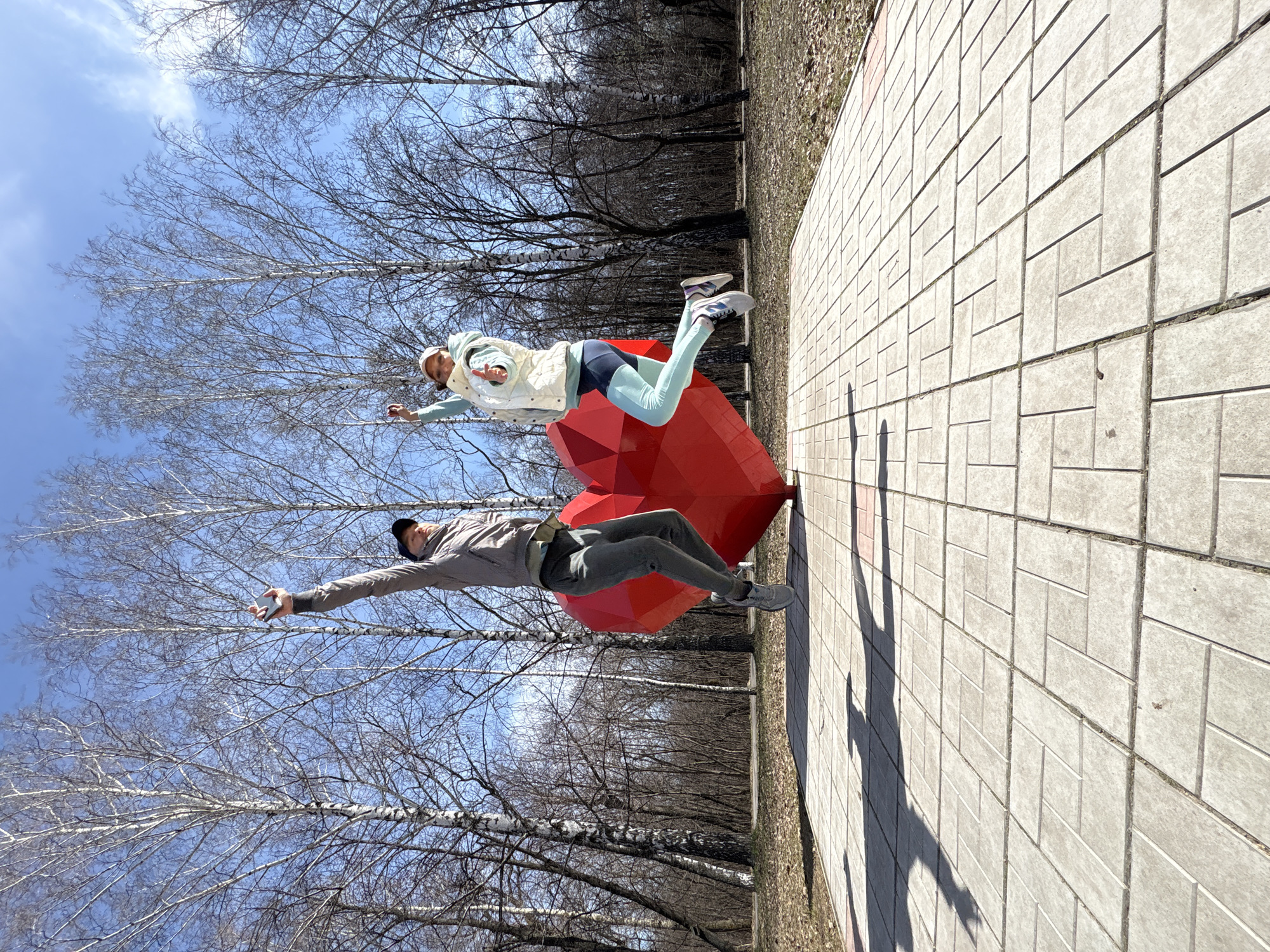 This image captures a playful outdoor scene featuring two people, a man and a woman, posed as if floating in mid-air next to a large, red, geometric heart sculpture. The photo is set in what appears to be a park or public area in Ulyanovsk, Russia, indicated by the bare birch trees characteristic of early spring or late autumn in that region.
The woman, positioned to the right of the heart sculpture, wears a light-colored vest over a long-sleeved shirt, light blue leggings, and athletic shoes, topped with a cap. Her arms are slightly outstretched, and her legs are bent, creating the illusion of weightlessness. The man, to the left of the sculpture, is dressed in a grey jacket, dark pants, and athletic shoes, with a dark cap. He mirrors the woman's pose, extending his arms and bending his legs as if suspended.
The prominent red heart sculpture, made of a multi-faceted material, is central to their playful interaction. The ground is a light grey paved pathway, composed of rectangular pavers laid in a repeating pattern. Long, distinct shadows of the two individuals stretch across the pavement, suggesting bright sunshine and either morning or late afternoon light. The background is filled with a dense array of bare tree branches against a clear blue sky with a few wispy clouds. The overall mood conveyed is lighthearted and joyful. There is no visible text in the image.