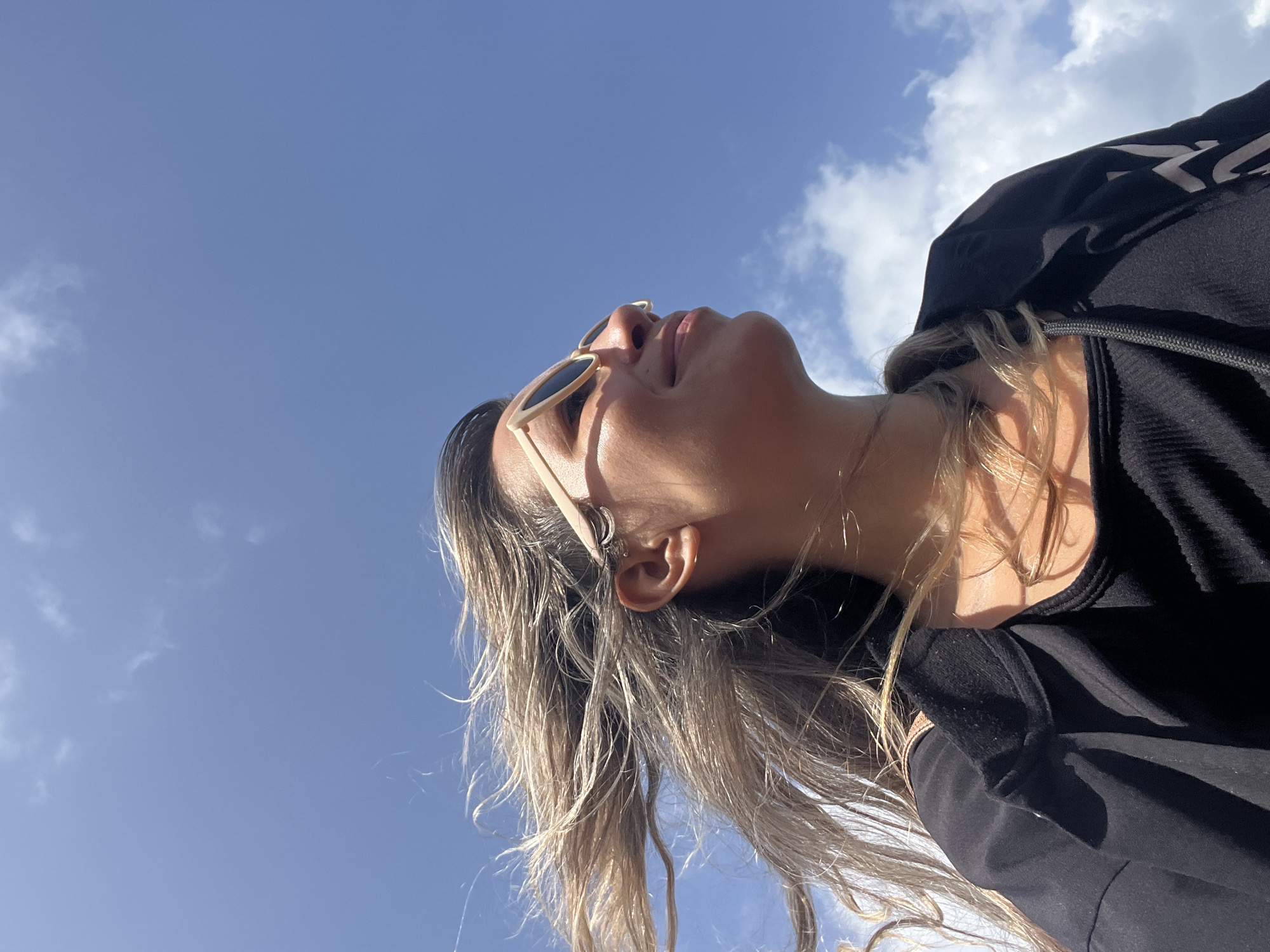 A medium shot shows a person's head and shoulders from a low angle, with the sky dominating the background. The person, facing to the right, has long, windblown blonde hair and is wearing light-colored sunglasses and a black jacket over a black top. The person's face is partially visible, and they appear to be smiling. The sky is blue with scattered white clouds. The image was taken in Walgama, Sri Lanka.