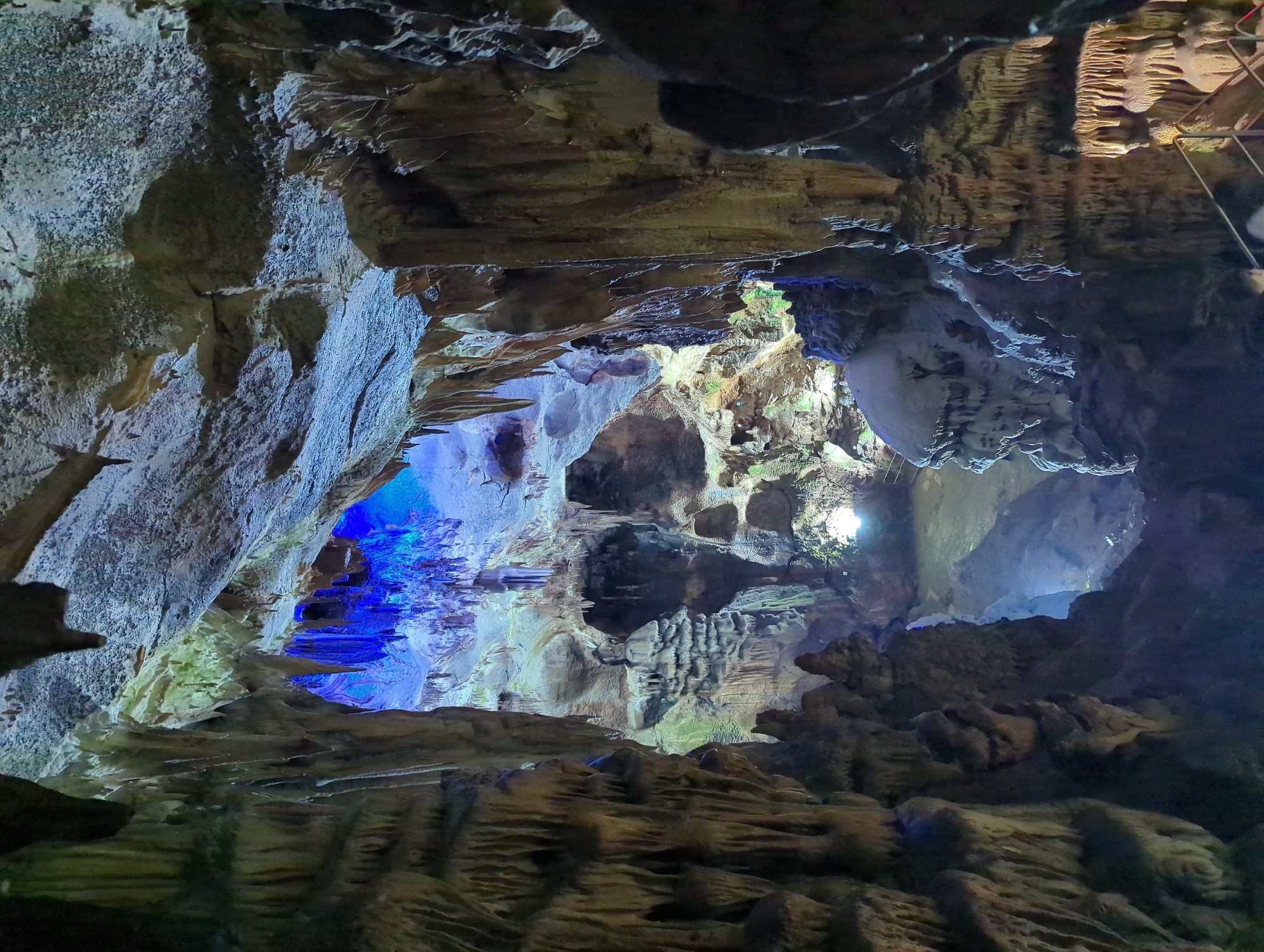 This image captures the interior of the Ledenika Cave, a natural wonder located in the Vratsa Balkan Mountains, near the city of Vratsa, Bulgaria. The cave's formations are illuminated by artificial lighting, casting a blue glow on the stalactites and stalagmites. The foreground is dominated by textured cave walls and numerous stalactites hanging from the ceiling, some appearing as thin, sharp icicles, while others are thicker and more rounded. In the mid-ground, the cave opens up to reveal a more expansive cavern with layered rock formations and a pool of water reflecting the blue light. Deeper in the cave, more intricate stalactite and stalagmite structures are visible, sculpted by millennia of water erosion. The ceiling is a complex tapestry of rock formations, some smooth and others rough, with the blue light creating a dramatic and ethereal atmosphere. The lighting also accentuates the mineral deposits on the cave walls, giving them a textured, almost crystalline appearance. The overall scene is one of natural grandeur, enhanced by strategic illumination to highlight the geological features.