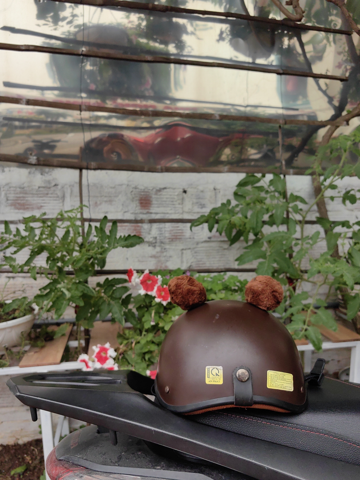 A brown helmet with two fuzzy brown balls attached to the top sits on the black seat of a motorcycle. The helmet has a black chin strap and two small rectangular stickers on the back. Behind the motorcycle, there is a small garden with green plants and red and white flowers. The background includes a wall with horizontal white lines, partially covered by reflective material that distorts the image behind it. Based on the architectural style and foliage visible in the image, the location is likely in Bai Chay Ward, Vietnam.