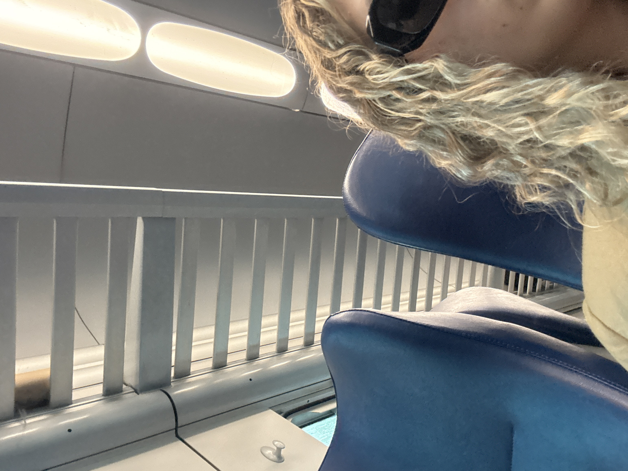The image shows a person with blonde, curly hair wearing dark glasses, sitting in a blue seat on what appears to be a train or bus. A white railing with vertical bars is visible in the background, next to a white wall. There are two bright, elongated lights on the ceiling. There is no verifiable information present in the image to confirm the specific location, Alessandria, Italy.