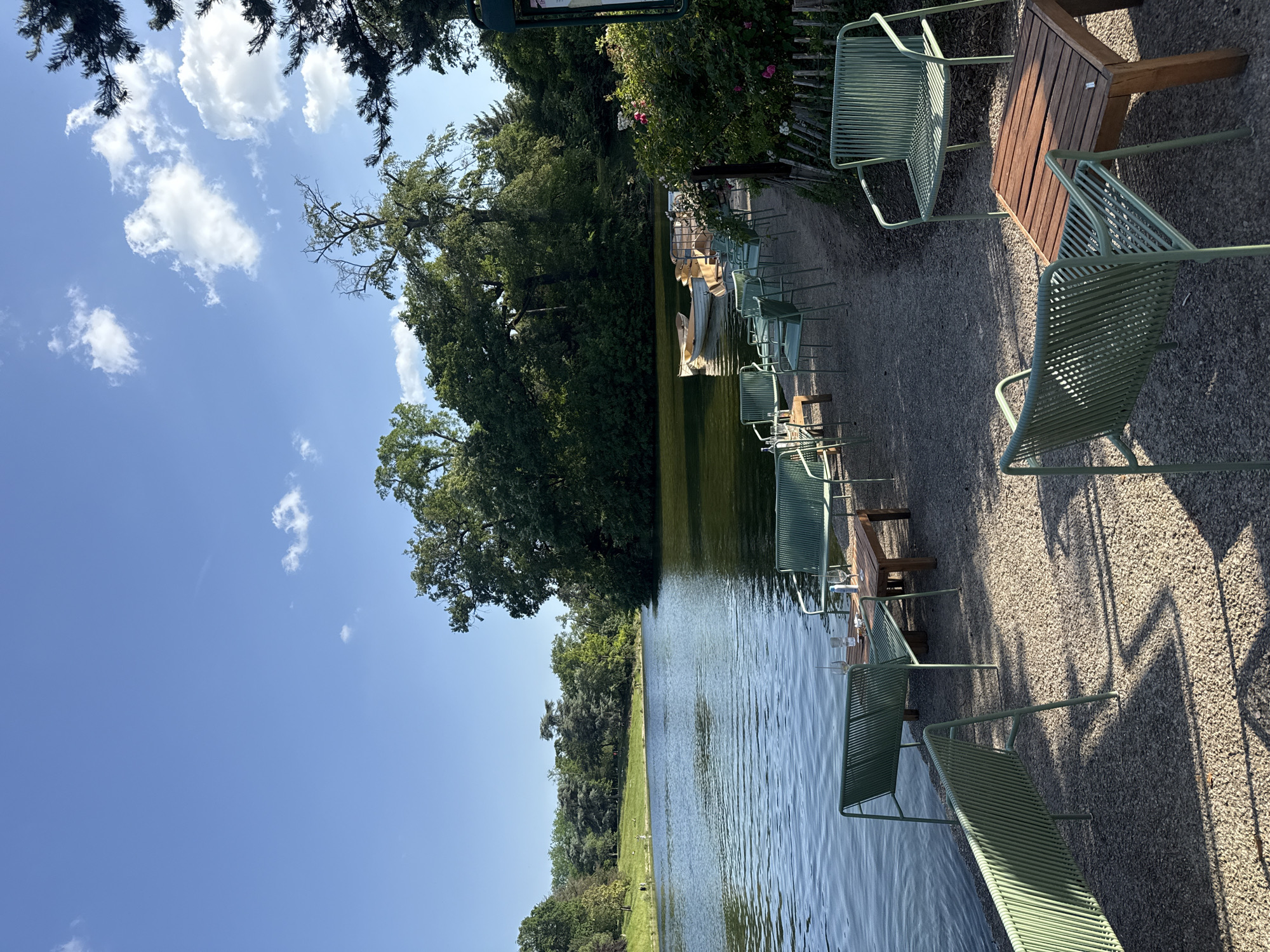 This image depicts a scenic view of a body of water bordered by trees and a seating area with green metal chairs and wooden tables. The chairs are arranged along a gravel path next to the water. In the background, there are boats docked along the shore. The sky is blue with scattered clouds. This scene takes place in Paris, France.