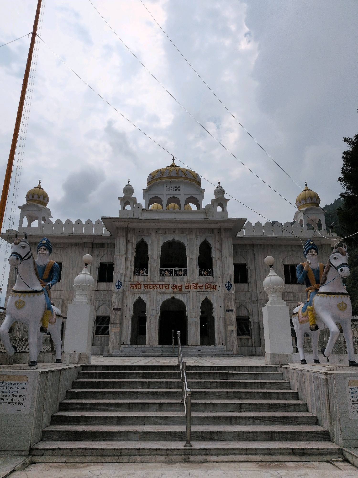 The image depicts the entrance to the Gurdwara Gobind Singh in the city of Mandi, India. The Gurdwara is a white marble building with golden domes, flanked by two large statues of men on white horses. The statues are dressed in traditional Sikh attire, including turbans and long tunics. The building has a series of arched entrances and windows, and a large flagpole stands to the left of the structure. A set of marble stairs leads up to the main entrance, with a metal handrail in the center. The sky is overcast with grey clouds.