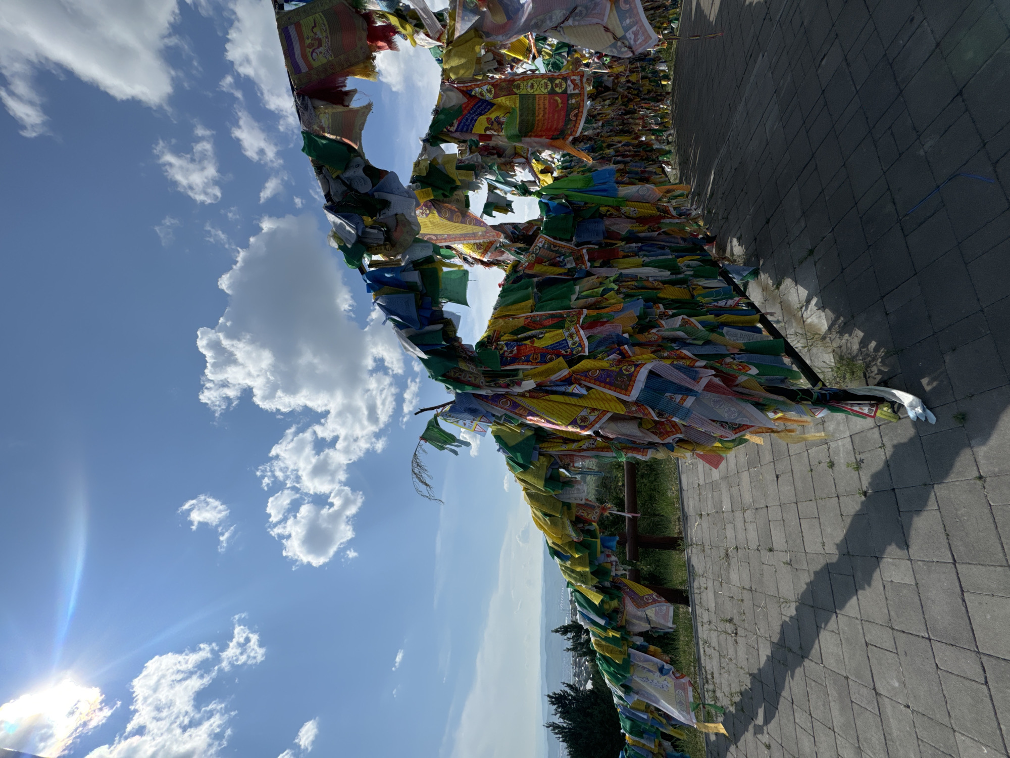 This is a wide-angle, low-angle, outdoor, daytime shot of a landscape with Buddhist prayer flags. The flags are predominantly yellow, blue, red, green, and white, strung across metal poles and wires. The flags are densely packed and appear weathered, with some showing wear and tear. They are arranged in a curvilinear fashion, creating a sense of depth and movement.
In the background, a hazy city skyline is visible, suggesting an urban environment. The city is framed by rolling hills and a lush green landscape with scattered trees. The sky is a vibrant blue, filled with fluffy white cumulus clouds. Sunlight streams down from the upper left corner of the image, creating lens flare and casting long shadows from the prayer flags onto the paved ground in the foreground. The ground is made of gray interlocking pavers.
The location is identified as a viewpoint on Lysaya Gora (Bald Mountain) in Ulan-Ude, Russia. This viewpoint is a popular spot known for its panoramic views of the city and is adorned with numerous prayer flags. The scene depicts a place of spiritual significance and natural beauty. There are no people visible in the image.