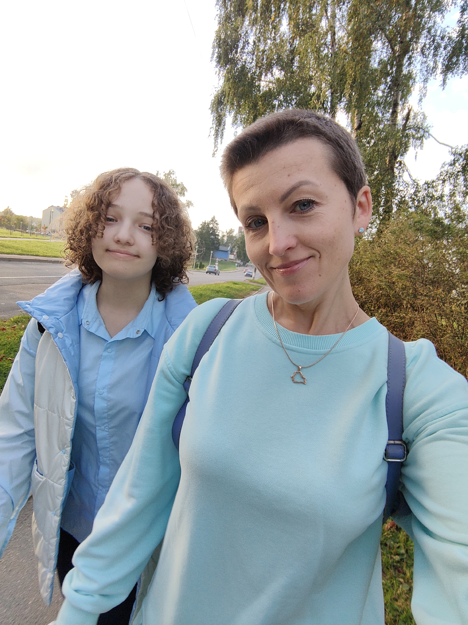 This is a selfie taken outdoors, likely on a sidewalk or path, with a paved road and grass visible in the background. Two individuals, a woman and a younger girl, are in the foreground. The woman, in the right of the frame, has short brown hair and is wearing a light blue sweatshirt and a necklace with a pendant. The girl, on the left, has curly brown hair and is wearing a light blue button-down shirt and a light-colored jacket. Trees and some buildings are visible in the distance, suggesting an urban or suburban environment. The lighting indicates it is daytime, possibly late afternoon due to the warm tone of the light. The specific location of Fanipal, Belarus is not visually discernible from the image itself.
