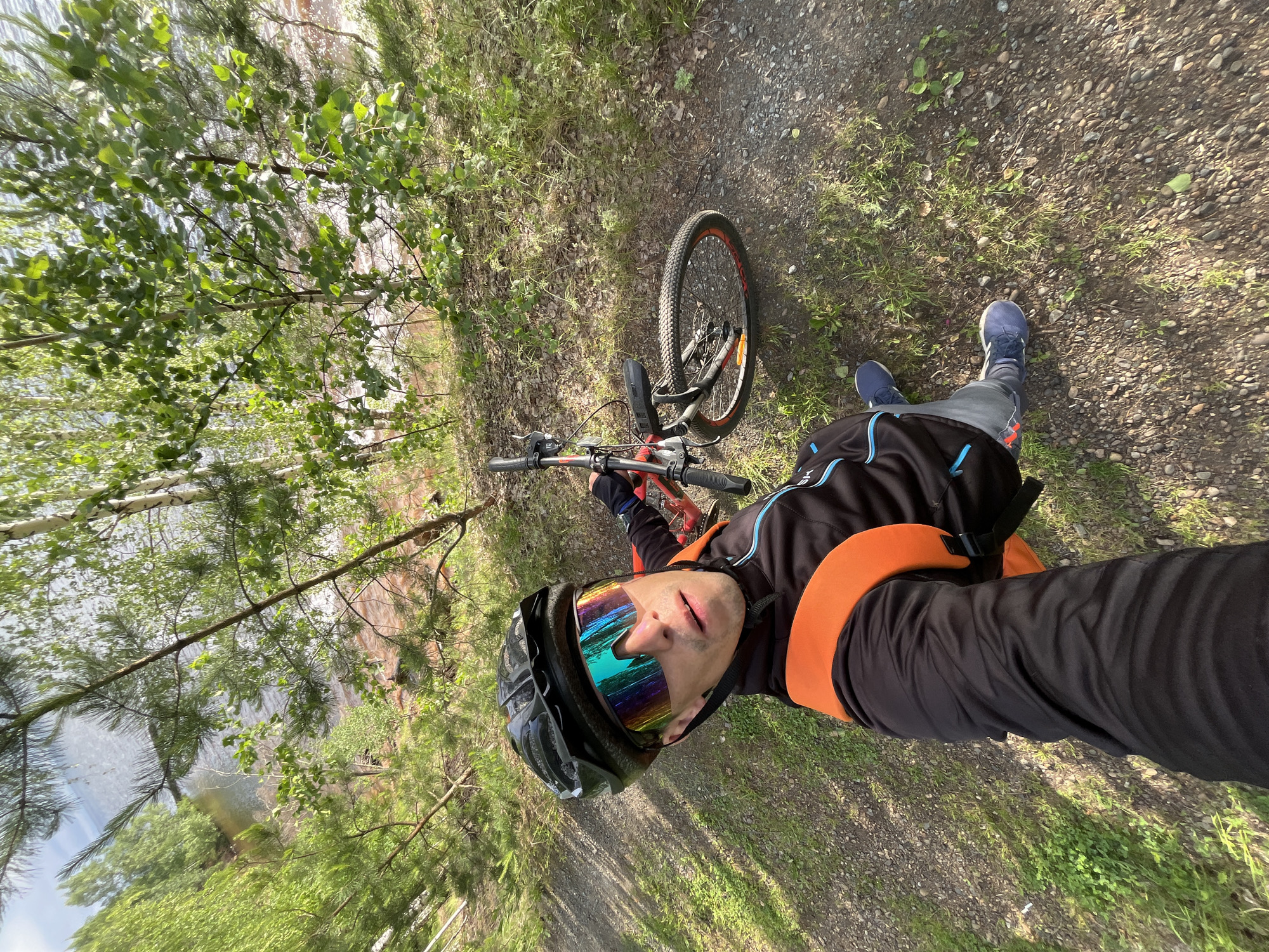 A man in a black helmet and mirrored cycling glasses is taking a selfie with his red mountain bike. He is wearing a black jacket with an orange vest and blue pants. The bike is leaning against a tree. The man is standing on a gravel path in a wooded area with a body of water visible in the background. The environment appears to be natural and outdoors.