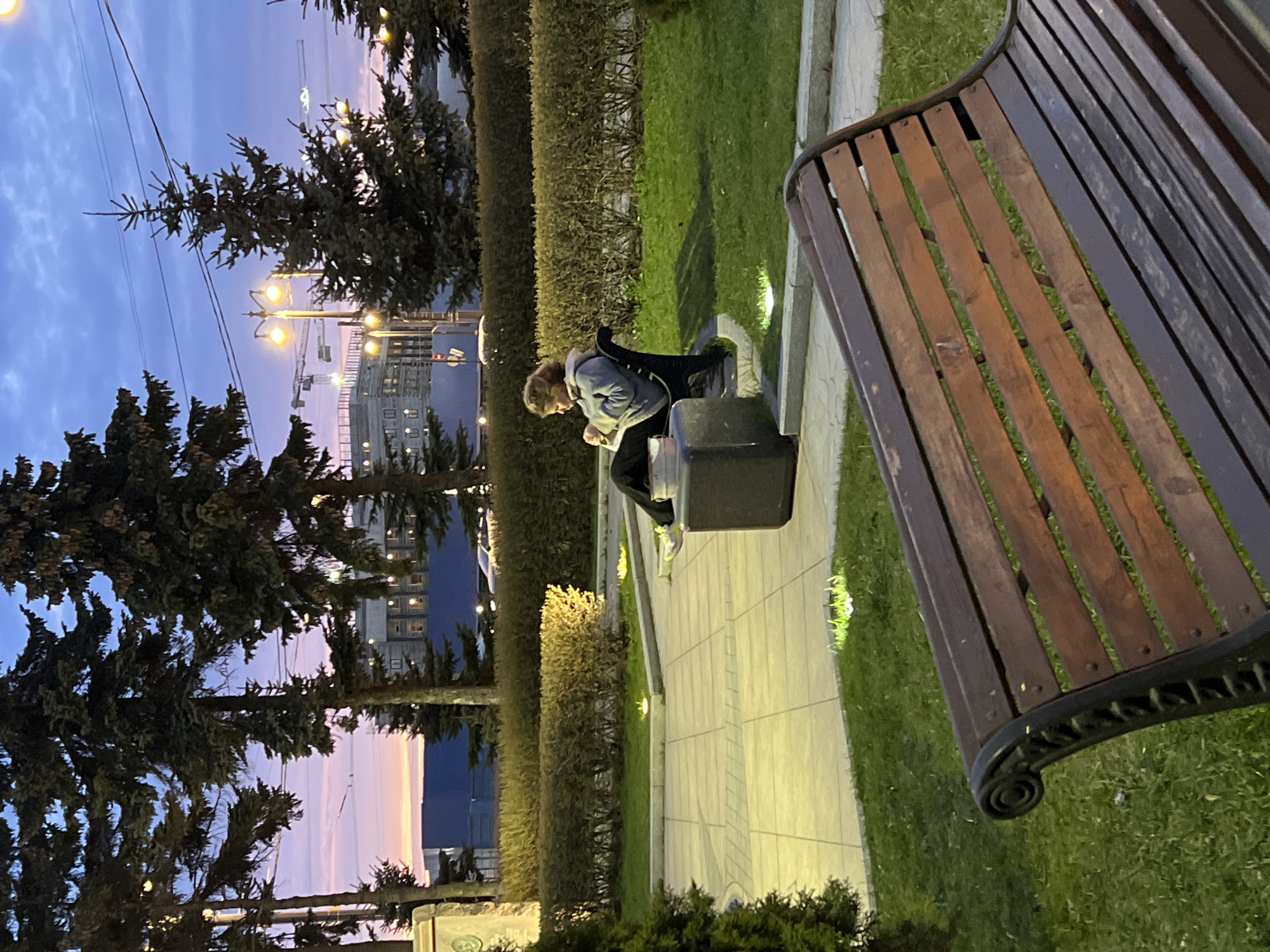 The image captures a park scene in Saint Petersburg, Russia, at dusk. A person with fair hair, clad in a gray jacket and dark pants, is seated on a dark-colored box. The person appears to be interacting with something held in their hands. A wooden park bench is visible in the foreground. The background shows a manicured hedge, trees, and a building illuminated by streetlights. The sky displays a gradient of colors from blue to orange, indicating twilight.
