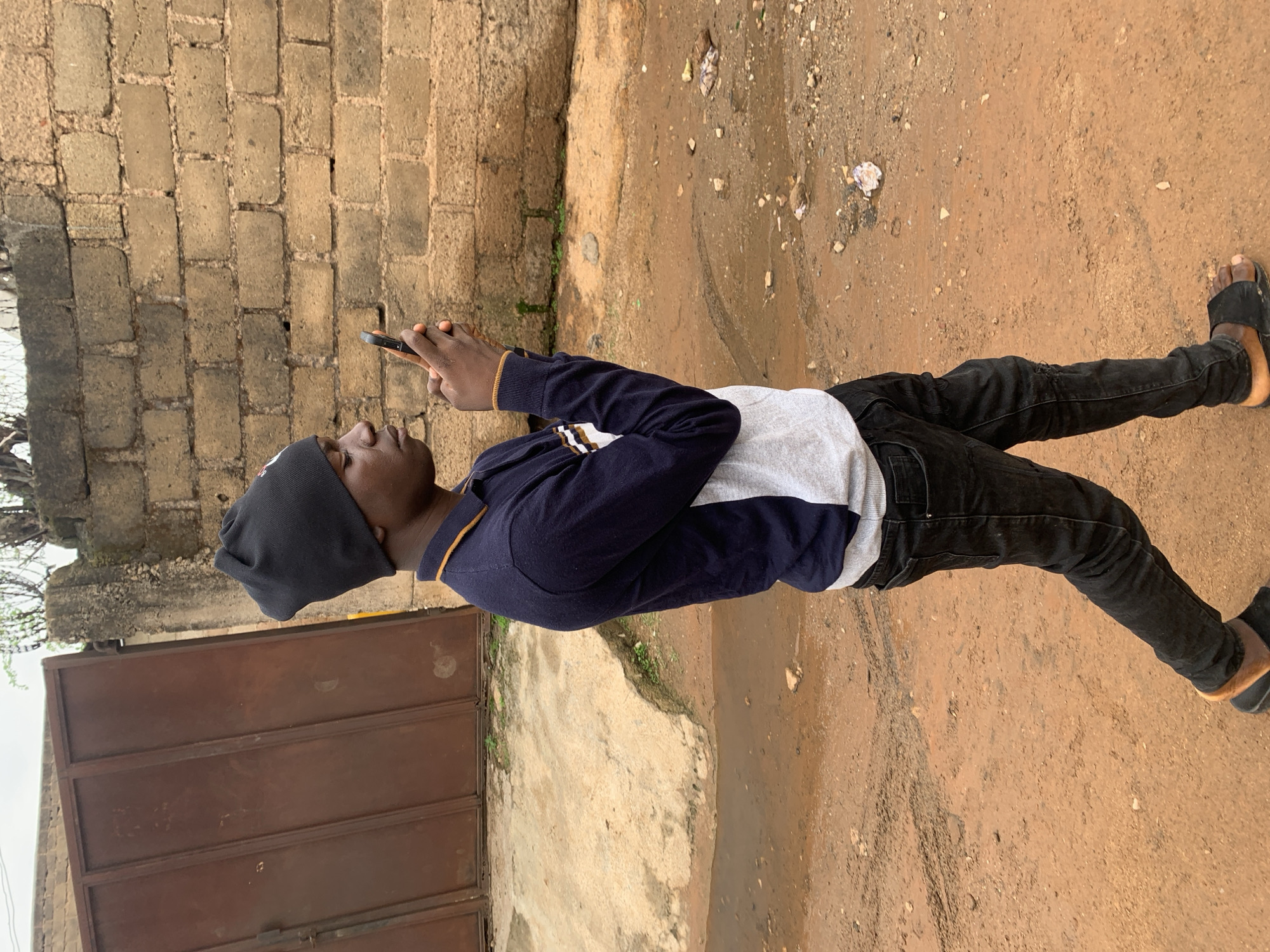 A dark-complexioned male subject, estimated to be a young adult, is depicted in profile, walking on an unpaved reddish-brown dirt path in Gura Top, Nigeria. He is dressed in a black beanie, a navy blue long-sleeved polo shirt featuring horizontal white and tan stripes across the chest and on the arm cuffs, black denim jeans, and black slide sandals with tan soles. The subject holds a dark-colored smartphone vertically with both hands, directing his gaze downwards at the device. His posture indicates movement, with the left foot on the ground and the right foot lifted.
The ground consists of uneven reddish-brown dirt, interspersed with small puddles and patches of moisture, particularly in the mid-ground. Scattered small rocks and light-colored debris are visible on the surface. To the left of the subject, a large, weathered brown metal gate composed of multiple vertical panels is present, set within a concrete wall structure. Behind the subject and extending to the right, a light brown block wall, showing signs of weathering and discoloration, forms the primary background. Sparse, dry-looking vegetation is visible above the wall line against an overcast sky.