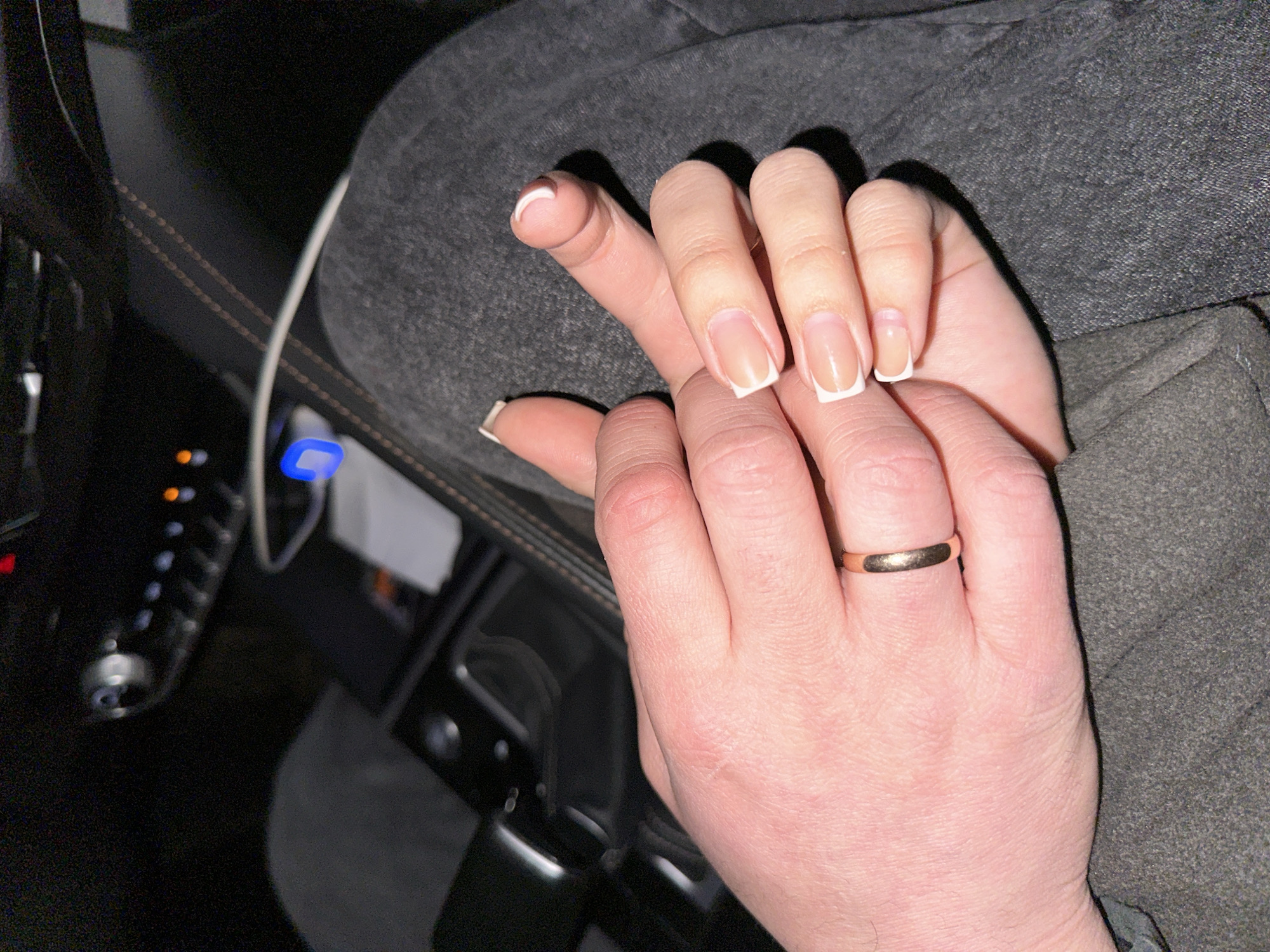 A close-up photograph shows two hands intertwined. The top hand, with manicured nails featuring a white French tip, rests on the lower hand, which is adorned with a simple gold ring on the ring finger. The setting appears to be inside a vehicle, evidenced by the car's interior components in the background, like the dashboard and a charging cable plugged into a device. The environment is dimly lit. The location in the city of Chaikovsky, Russia is not identifiable from this image.