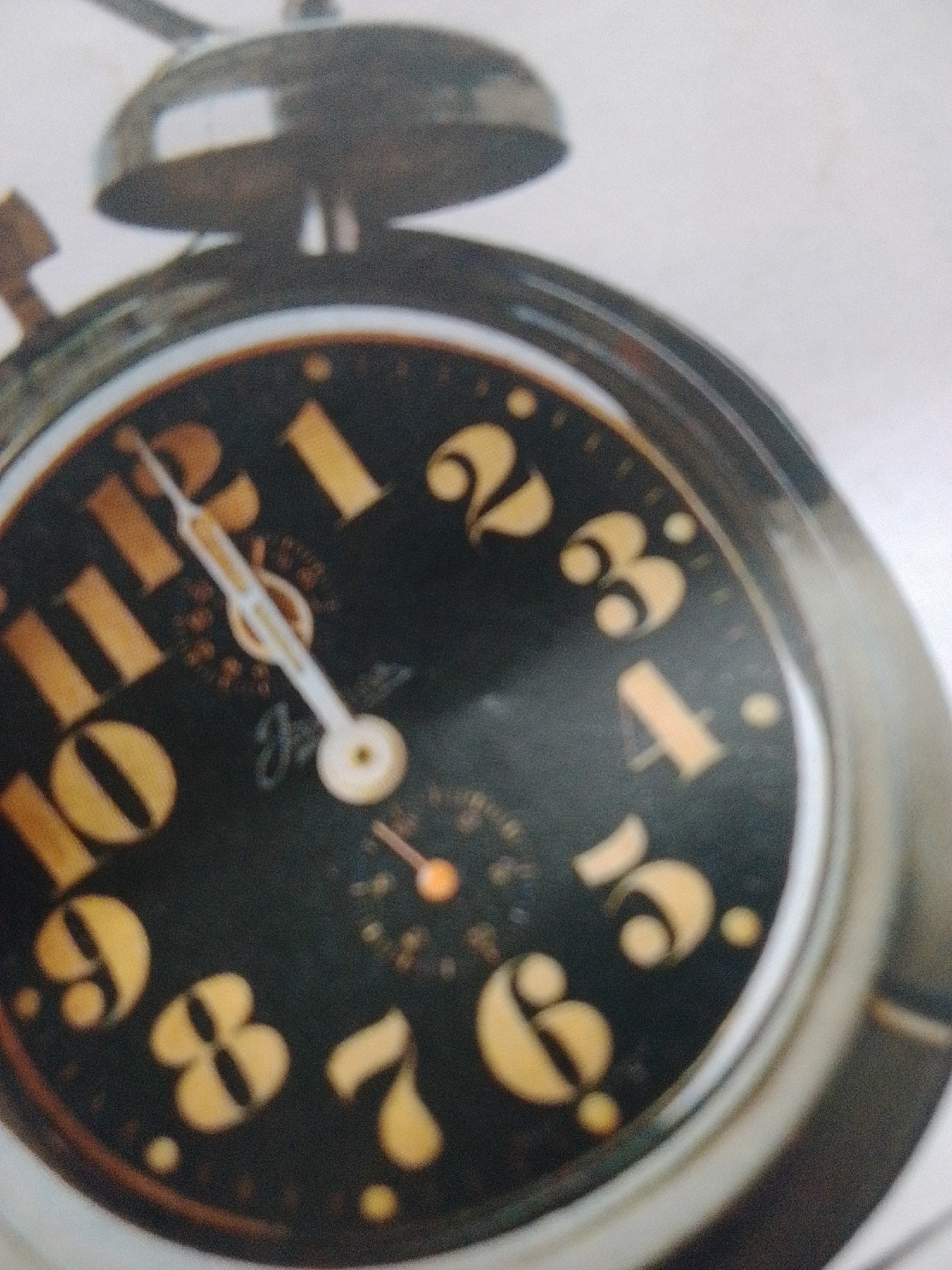 This is a close-up, slightly blurred image of an alarm clock with a black face and yellow numerals. The clock is tilted, with the top of the bell visible. The hands of the clock are white, with the hour hand pointing between the 11 and 12, and the minute hand pointing towards the 12. There is a smaller dial in the center of the clock face. The overall impression is that of an old-fashioned mechanical alarm clock. No people are visible, and the setting appears to be indoors due to the lighting. There is no visible text on the clock face other than the numerals. The lighting suggests it could be daytime.