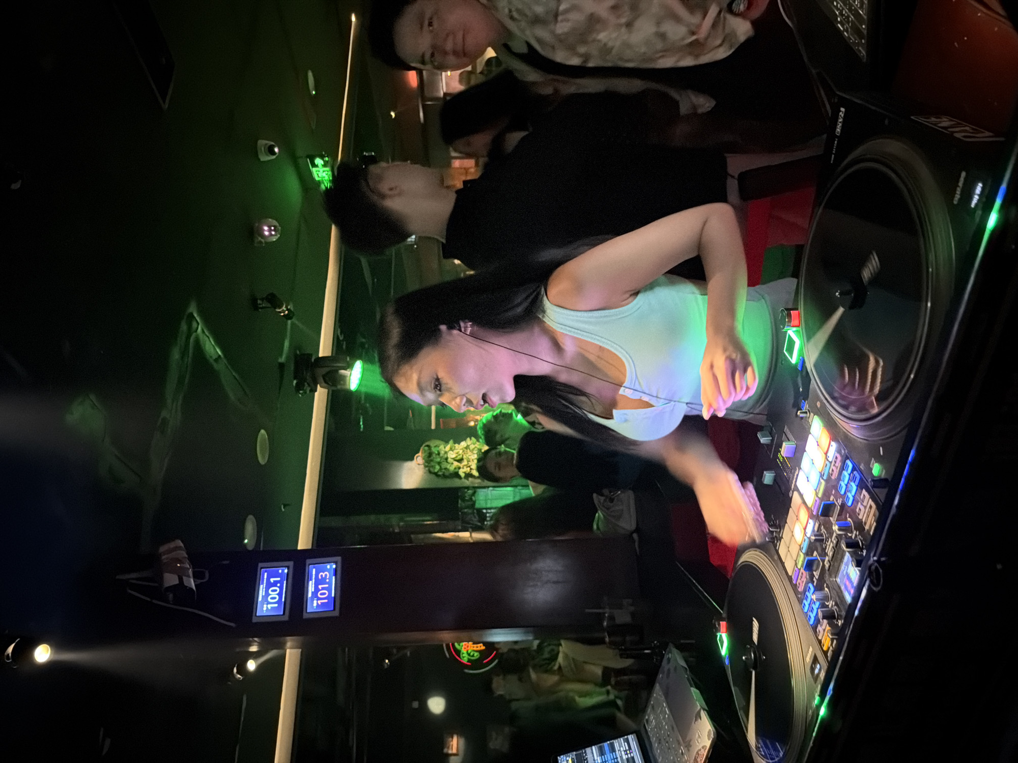 A female DJ, wearing a light blue tank top, is actively mixing music on a DJ console in a dimly lit club. She has long, dark hair and is wearing earbuds. Her hands are blurred from movement as she manipulates the controls. The DJ console features multiple illuminated buttons and two turntables. In the background, other patrons are visible, some standing and talking. The environment includes green accent lighting on the ceiling and structural elements with digital displays showing "100.1" and "101.3". The scene appears to be a nightclub in Liangjiang, China.