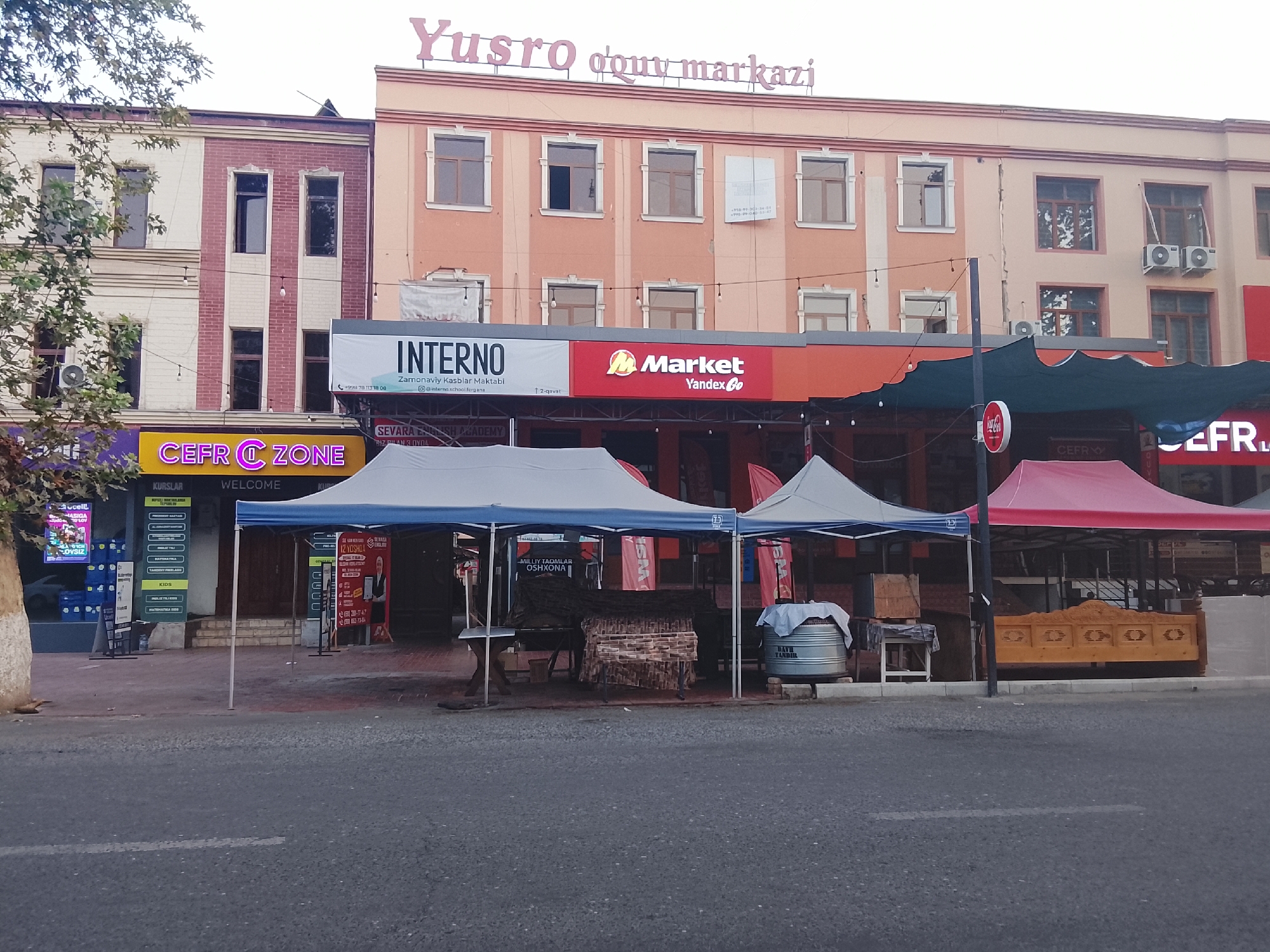 The image shows a street scene in Fergana City, Uzbekistan, with a multi-story building as the prominent backdrop. The building features a light orange facade with several rows of windows. Above the windows, a pink sign reads "Yusro qoviy markazi." Below this, a banner is visible with "INTERNO" and "M Market Yandex Go" logos. Several stalls with blue and red awnings are set up in front of the building, displaying various items. A street runs through the foreground of the image.