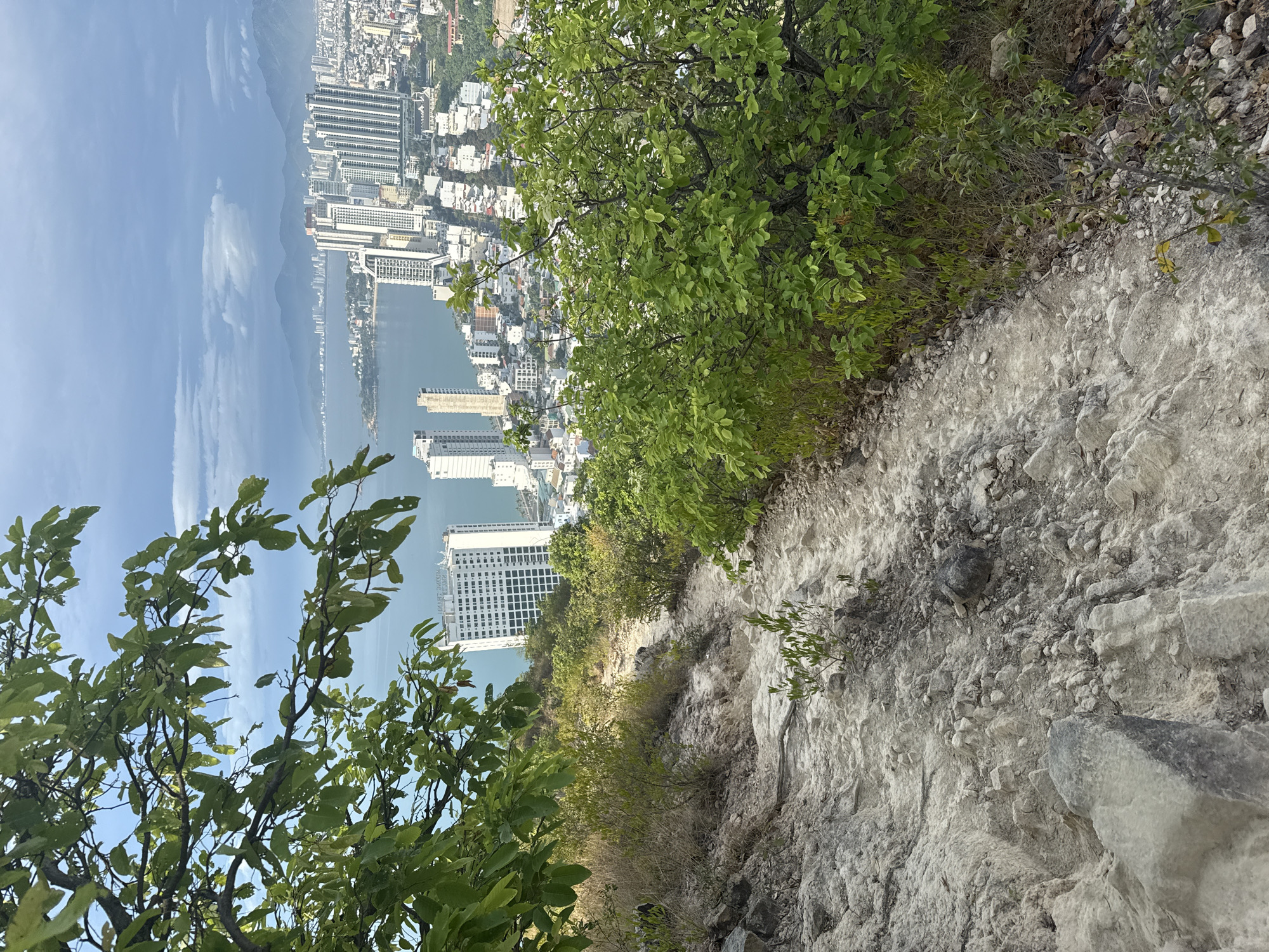 The image shows a downward view of a rocky, narrow path leading towards the coastal city of Nha Trang, Vietnam. The path is bordered by vegetation, with trees and shrubs framing the view of the city below. The city consists of tall buildings, the coastline, and a body of water, likely the sea or a bay. The sky is partially visible with some cloud cover. The photograph seems to have been taken from a high vantage point on a slope or cliff.