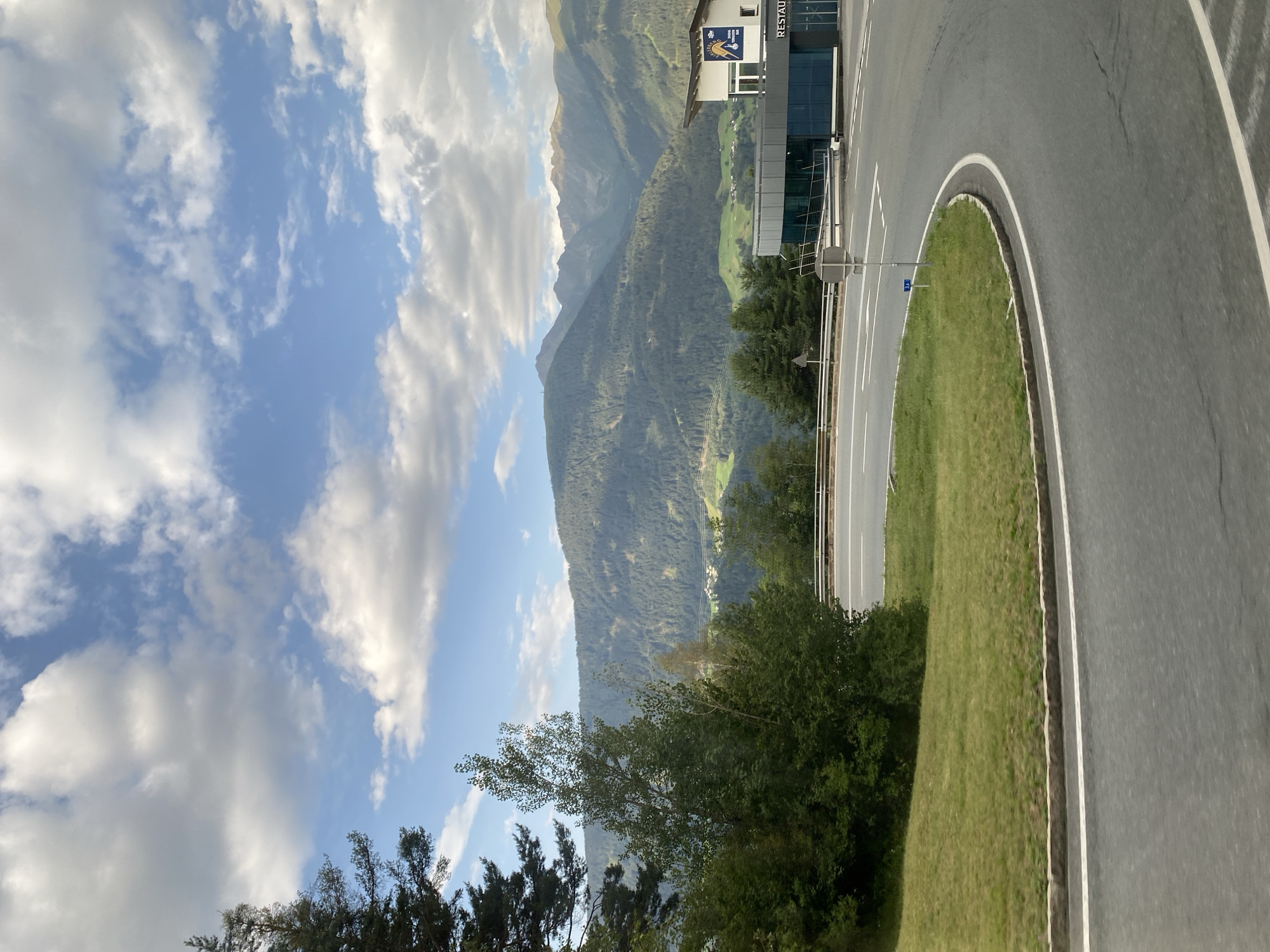 The image shows a curved road in a mountainous region with a building that appears to be a restaurant. The road is asphalt with white lane markings and a grassy median. In the background, there are dense green forests covering the slopes of large mountains, and a village is visible in the valley. The sky is blue with scattered white clouds. The building has a modern design with large glass windows and a prominent sign that reads "RESTAURANT". A conical evergreen tree is visible on the left side of the image, and other deciduous trees are present. This scene is located in the vicinity of Zirl, Austria.