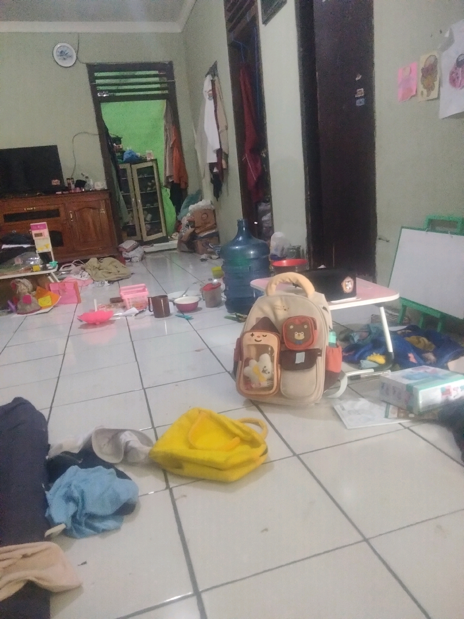 This image depicts a cluttered interior scene, likely a living space or play area, with white tiled flooring. Various items are scattered across the floor, including clothing, bags, bowls, and a large blue water jug. In the foreground, a tan backpack with cartoon animal designs is prominent. To the left, a wooden entertainment center is visible with a television on top. A doorway leading to another room with green walls is also present, revealing more items stored in cabinets. The overall impression is one of disarray, suggesting a recently used or untidy environment. The scene appears to be located in Suka Sari, Indonesia, based on the provided context.