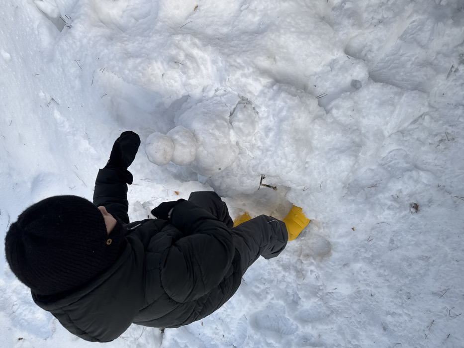 In Chaikovsky, Russia, a single individual is depicted outdoors, seen from an overhead perspective, engaged in building a snowman on snow-covered ground. The person wears a black ribbed knit beanie, a black puffer jacket with a hood, black gloves or mittens, dark-colored snow pants, and bright yellow boots. Their right arm is extended forward, positioned near a two-tiered snowman consisting of two spherical snowballs stacked vertically, the upper one smaller than the lower. The ground is entirely covered in white snow, exhibiting some compaction and scattered dark debris, possibly pine needles or twigs. A small, dark, T-shaped object is partially embedded in the snow near the individual's right foot.