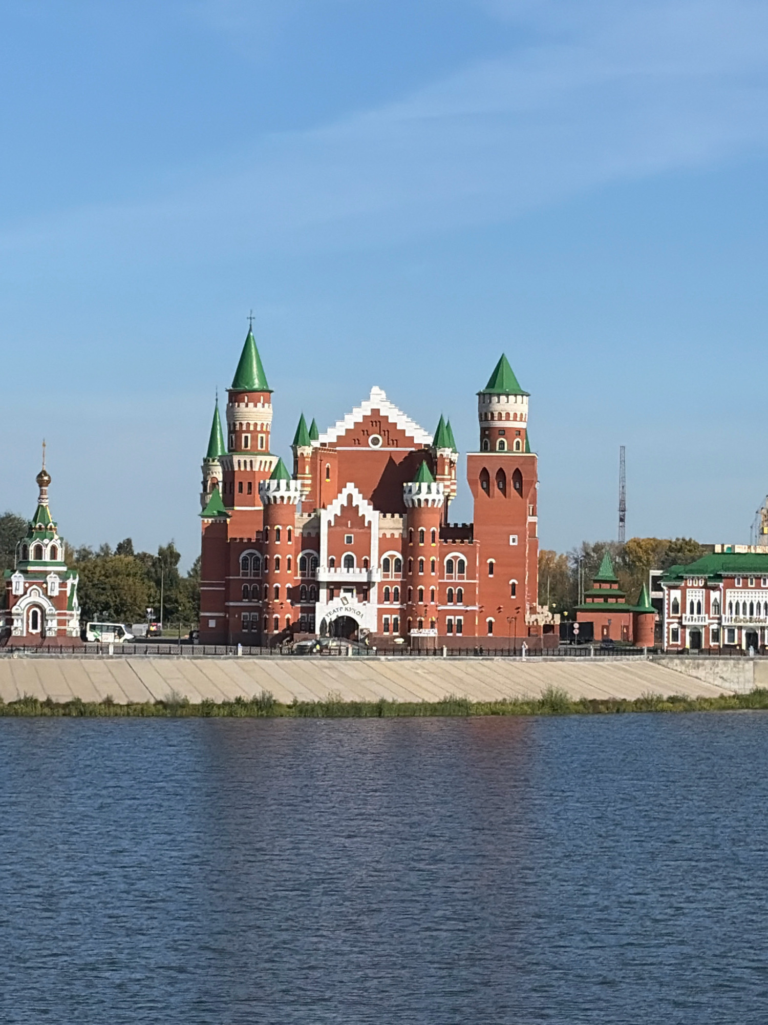 This image depicts a section of the embankment along the Malaya Kokshaga River in Yoshkar-Ola, Russia. The prominent architectural feature is the National Puppet Theatre, designed to resemble a medieval castle with red brick walls and green conical roofs. To the left of the theater, a smaller, ornate Orthodox church with green and white accents and a golden dome is visible. The embankment itself is lined with a concrete wall and a paved walkway, with trees and some vegetation visible behind the structures. Several vehicles, including a white bus, are present on the walkway. The sky is clear and blue.