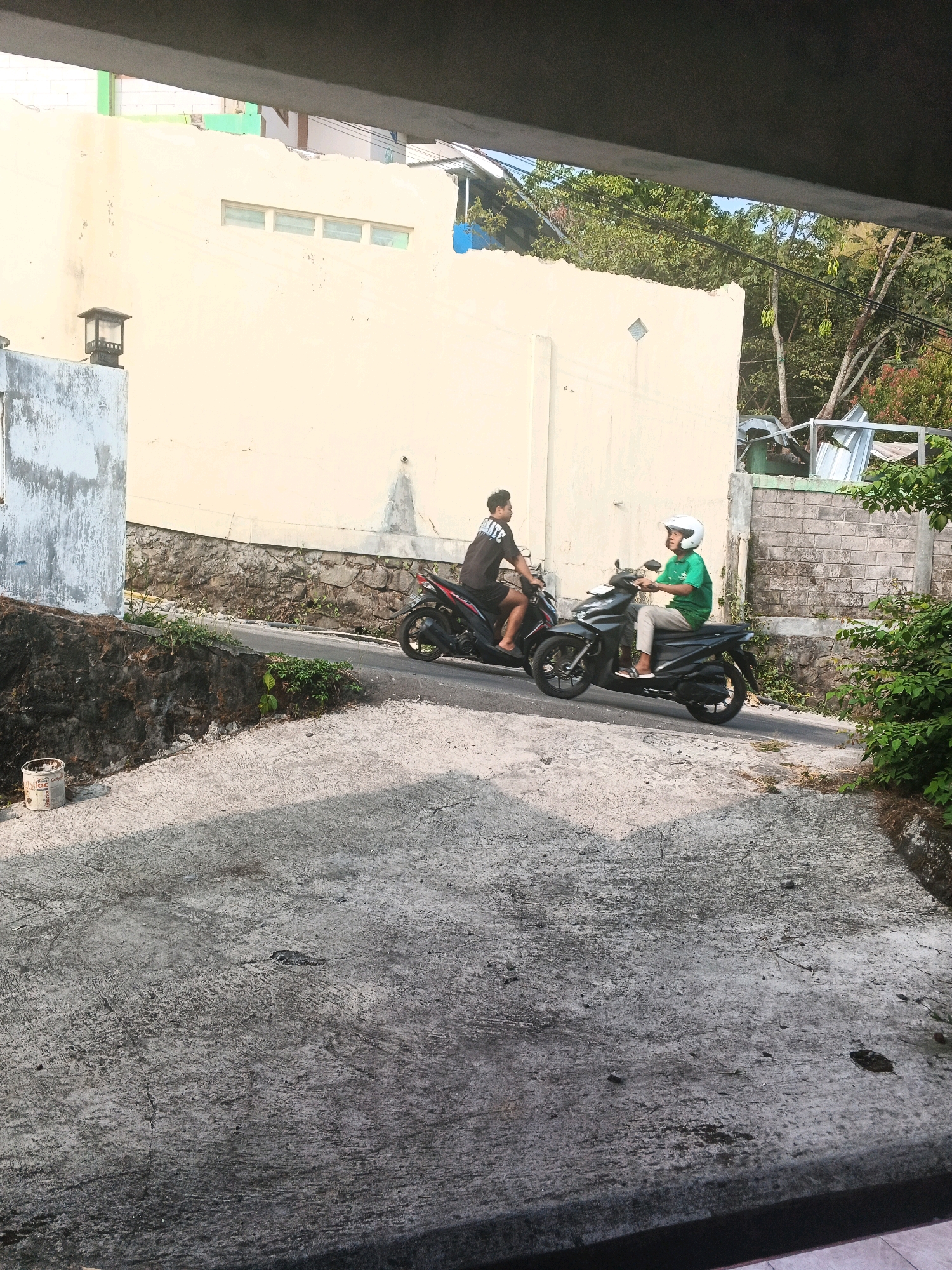 In Keji, Indonesia, two individuals are captured on scooters on a paved street. The first scooter, a black automatic model, is driven by a person wearing a black t-shirt and shorts. Behind them, a second black automatic scooter is ridden by an individual in a green t-shirt and light-colored pants, wearing a white helmet. The street is bordered by a stone retaining wall on the left and a yellow building with windows on the right. Trees and other vegetation are visible in the background, with a clear sky overhead. The foreground features a rough, grey concrete surface, possibly a driveway or sidewalk, with some debris scattered on it. A black lantern-style light fixture is mounted on a grey pillar to the left of the street.