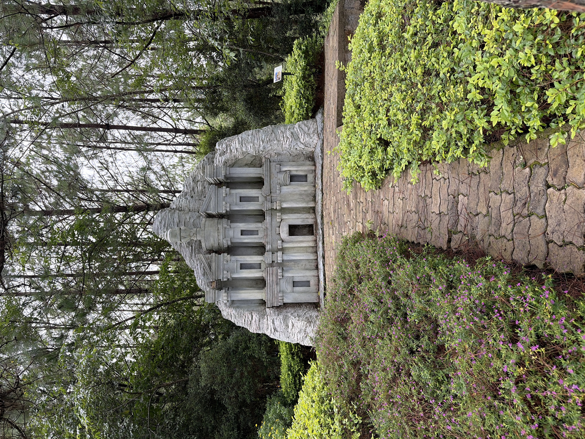 In the city of Lâm Viên - Đà Lạt, Vietnam, a miniature replica of a classical building stands amidst a lush, green park. The structure, resembling an ancient temple or amphitheater, is crafted from light grey stone and features multiple columns, arches, and recessed windows. It is set against a backdrop of tall, slender pine trees and dense foliage, suggesting an elevated or mountainous location. A pathway made of interlocking stone pavers leads towards the replica, bordered by vibrant green bushes and clusters of small purple flowers on the left, and well-maintained green shrubbery on the right. A small informational sign is visible in the background, near the replica. The sky appears overcast, casting a soft, diffused light over the scene.