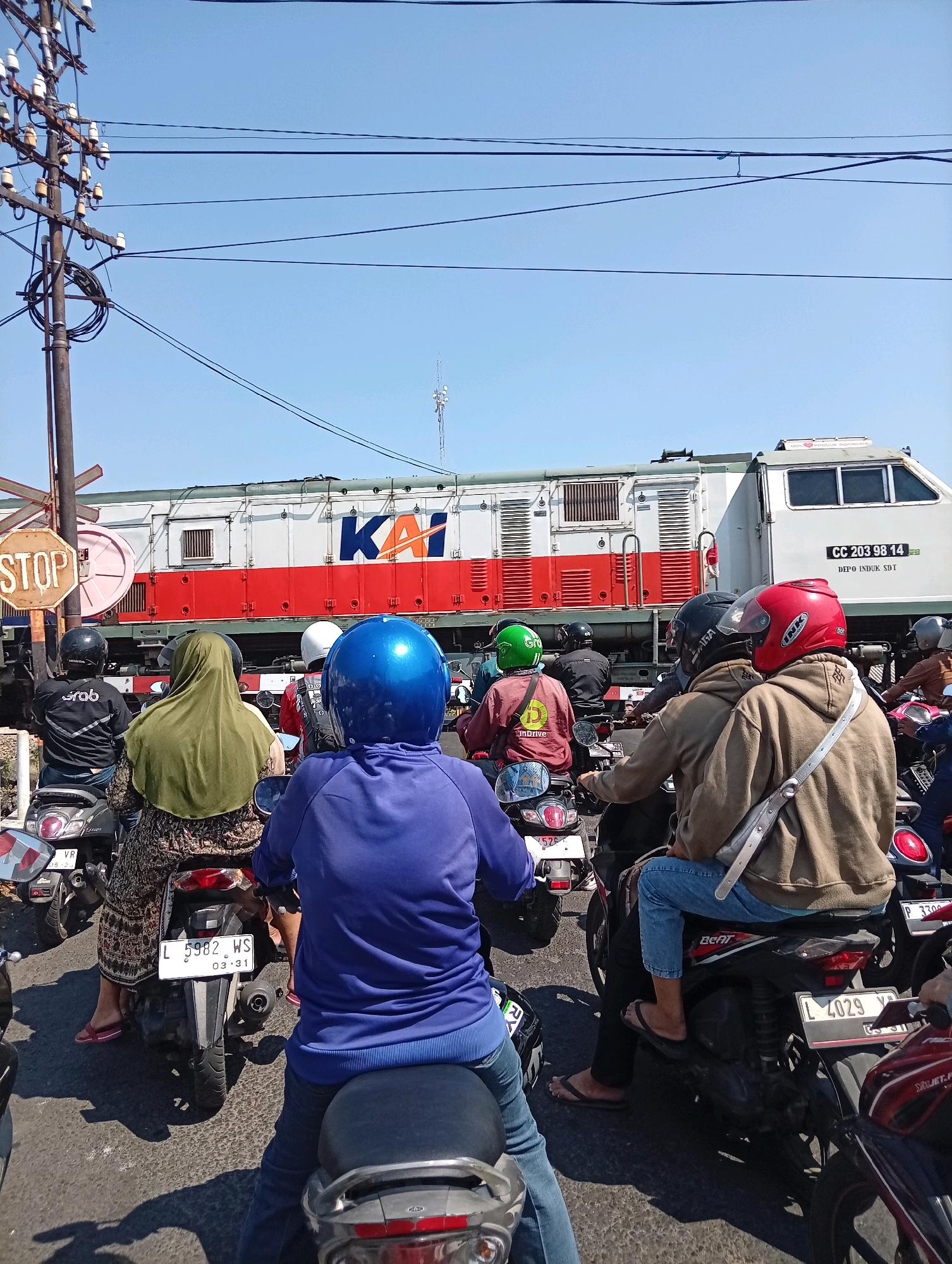This image captures a scene at a railway crossing in Surabaya, Indonesia, where a line of motorcycles is stopped, waiting for a train to pass. The train, emblazoned with the "KAI" logo, is a prominent subject, its red and white livery contrasting with the clear blue sky. Several individuals on motorcycles are visible, many wearing helmets in various colors, including blue, green, red, and black. Some riders are wearing casual clothing like hoodies and long skirts, with flip-flops also visible. The motorcycles are parked on a paved road, with shadows cast on the ground indicating a sunny day. A "STOP" sign is mounted on a pole to the left of the frame, further emphasizing the temporary halt. The presence of electrical poles and wires overhead suggests an urban or semi-urban environment. The license plates on the motorcycles, such as "L 5982 WS" and "L 4029 YX", are typical for Indonesian vehicles. The overall impression is one of everyday life interrupted by the passage of a train.