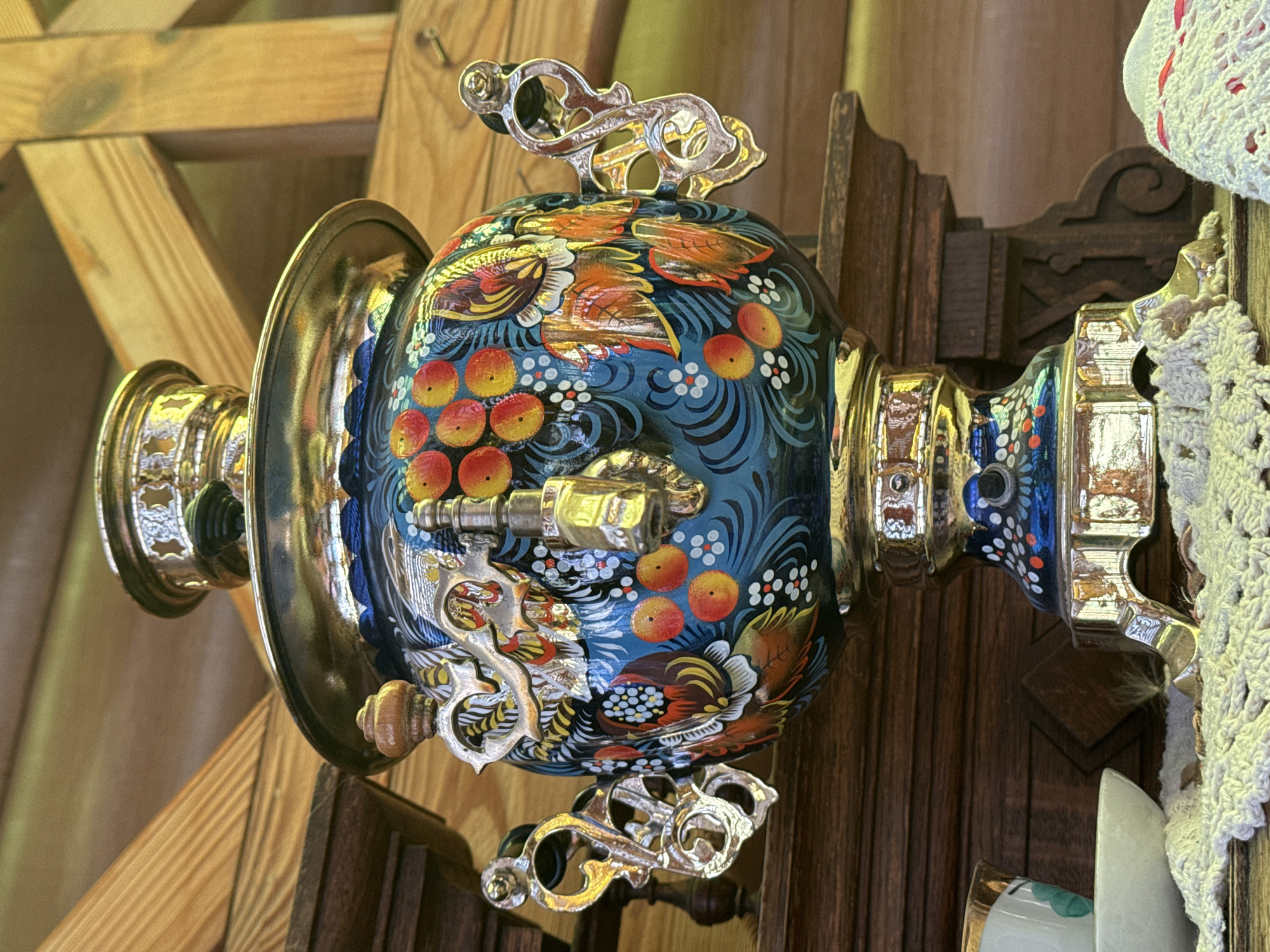 The image shows a Russian samovar, elaborately decorated with a blue background, red berries, and orange bird motifs. It features ornate silver handles and spigot. The samovar is positioned indoors, likely in a room with wooden walls and possibly part of a display or exhibition in the city of Kladovitsy, Russia. A white, crocheted object is visible on the right side of the image.