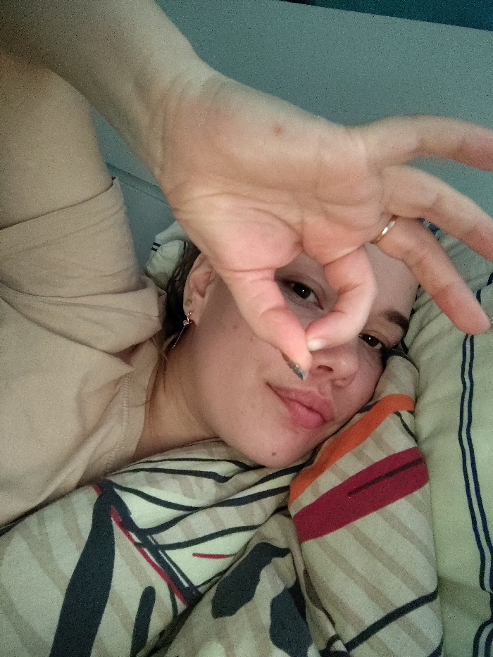 A young adult female is depicted in an indoor setting, presumably a bedroom within the city of Марковский, Russia. She is lying on her back, with her head resting on a pillow featuring green and white stripes. Her upper torso is visible, clad in a light beige garment. A patterned duvet, with horizontal and diagonal stripes in shades of beige, dark blue, red, and orange, covers her lower body. Her right arm is raised, with her hand positioned in front of her face, forming a circular shape with her thumb and index finger. Her left eye is visible through this opening, directly engaging the camera. A thin, gold-colored ring is present on the ring finger of her right hand, and dark nail polish is visible on her right thumb. A metallic earring with a dangling element is visible in her left ear. The background consists of a light blue wall or headboard.