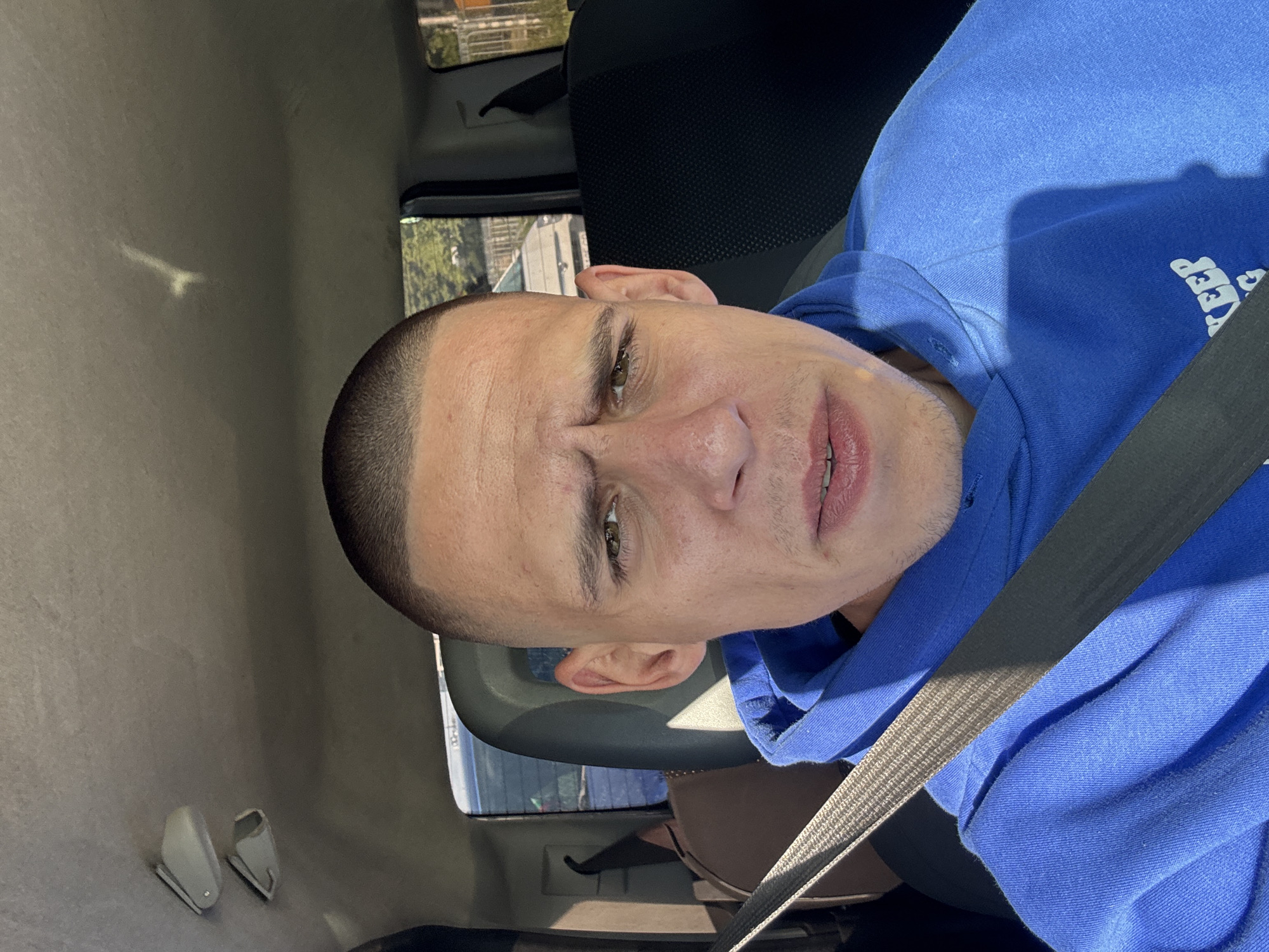 A man with a shaved head and wearing a blue hoodie is sitting in a car, wearing a seatbelt. The car appears to be moving, with a view of buildings and trees visible through the window. The location cannot be identified as Sofia, Bulgaria.