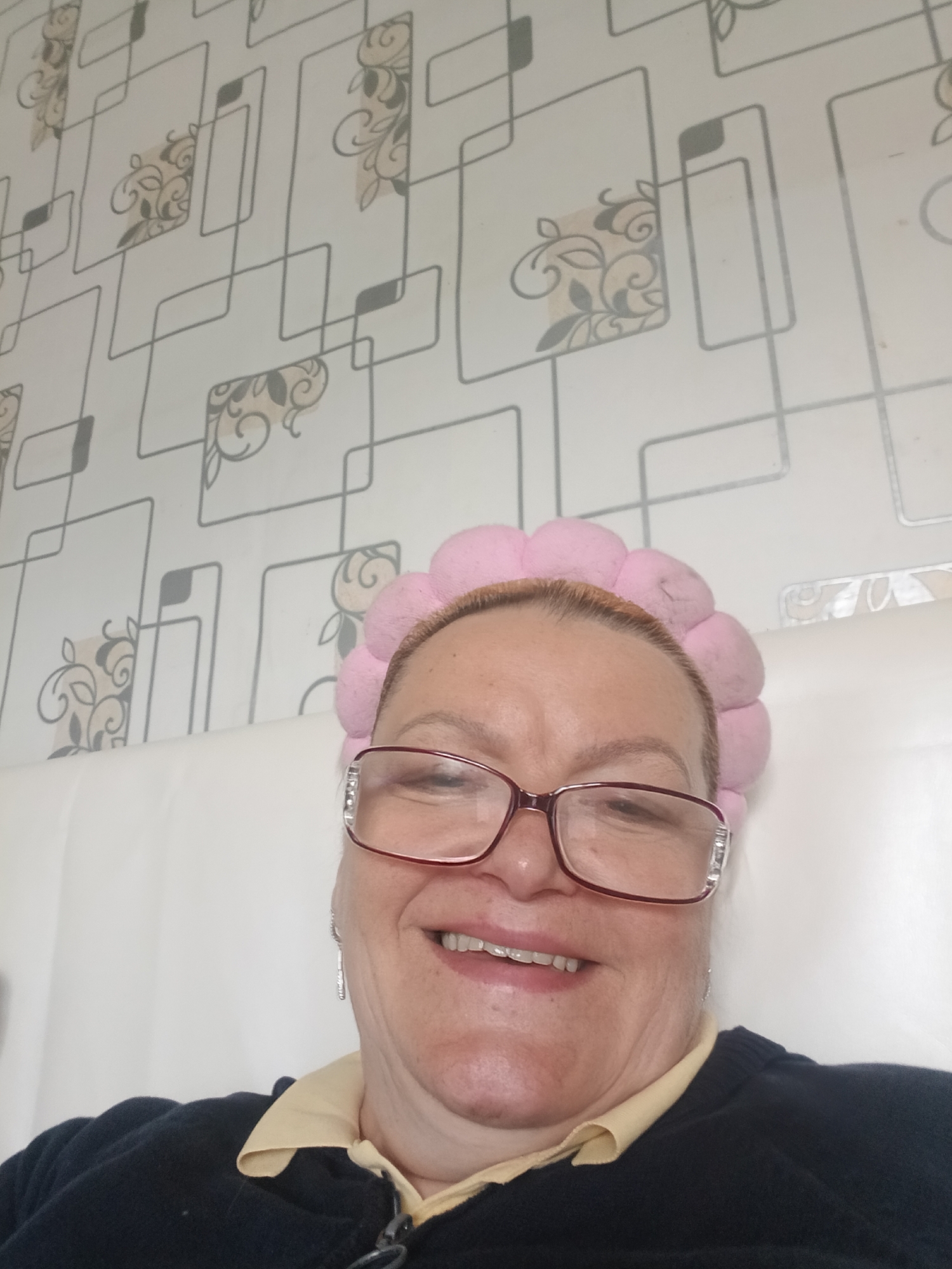 The image shows a selfie of a woman with reddish hair wearing a pink fluffy headband and red-rimmed glasses. She is smiling and wearing a dark blue sweater over a yellow collared shirt. Behind her is a white headboard and patterned wallpaper featuring gray lines forming squares with floral designs inside. There is no information to indicate the location is in Красный Маяк, Russia.