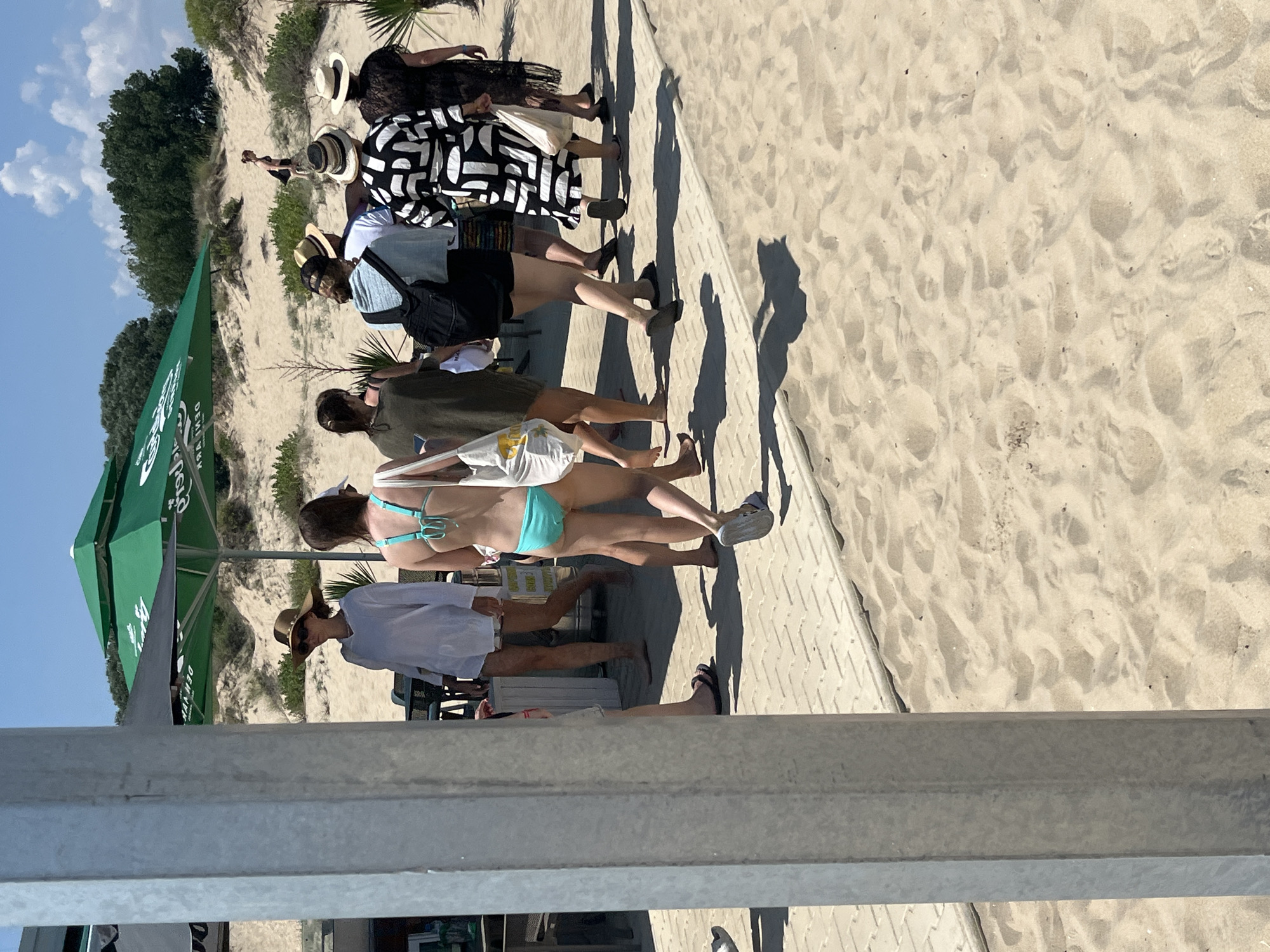 The image depicts a scene at a beach cafe in Nesebar, Bulgaria. Several people are walking on a paved pathway next to a sandy beach. In the background, a large green umbrella with visible text is providing shade. The pathway is made of light-colored rectangular stones, and the surrounding sand is textured with footprints.
In the foreground, a light gray metallic pillar is partially obscuring the view. To the left, a person's legs are visible, wearing dark flip-flops.
Moving towards the center and right of the image, a group of individuals are walking away from the camera.
The first person on the left is wearing a white cover-up and a wide-brimmed hat.
Next to them is a person in a turquoise bikini, carrying a white tote bag.
Behind them is someone in a dark green top and shorts, also carrying a white tote bag.
Further to the right, a person is wearing a black and white patterned dress and carrying a white tote bag.
To their right, another person is wearing a dark, possibly lace, dress.
Another individual in a straw hat and white shirt is visible in the background.
A person wearing a black swimsuit is seen walking up a sandy dune in the distance.
The environment is sunny with a bright blue sky and scattered white clouds. Lush green trees and bushes are visible on the dunes in the background. The overall impression is a casual summer day at a beachside establishment.