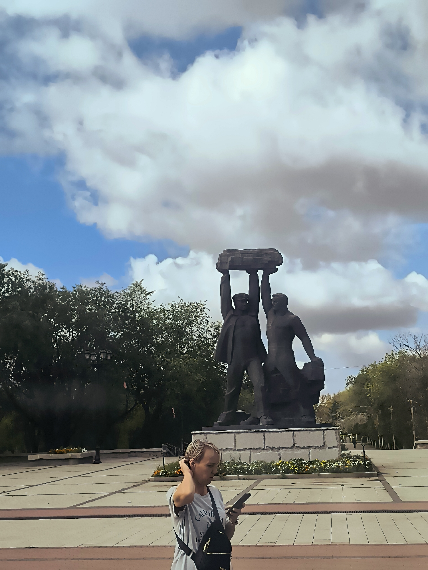 In Qarağandı, Kazakhstan, in front of the city administration building, a bronze sculpture depicts two male figures lifting a large, rectangular object overhead. The sculpture stands on a stone pedestal surrounded by a flower bed. In the foreground, a person with short blonde hair, wearing a white t-shirt and a backpack, is looking down at a smartphone. The background features trees and a paved public space under a partly cloudy sky.