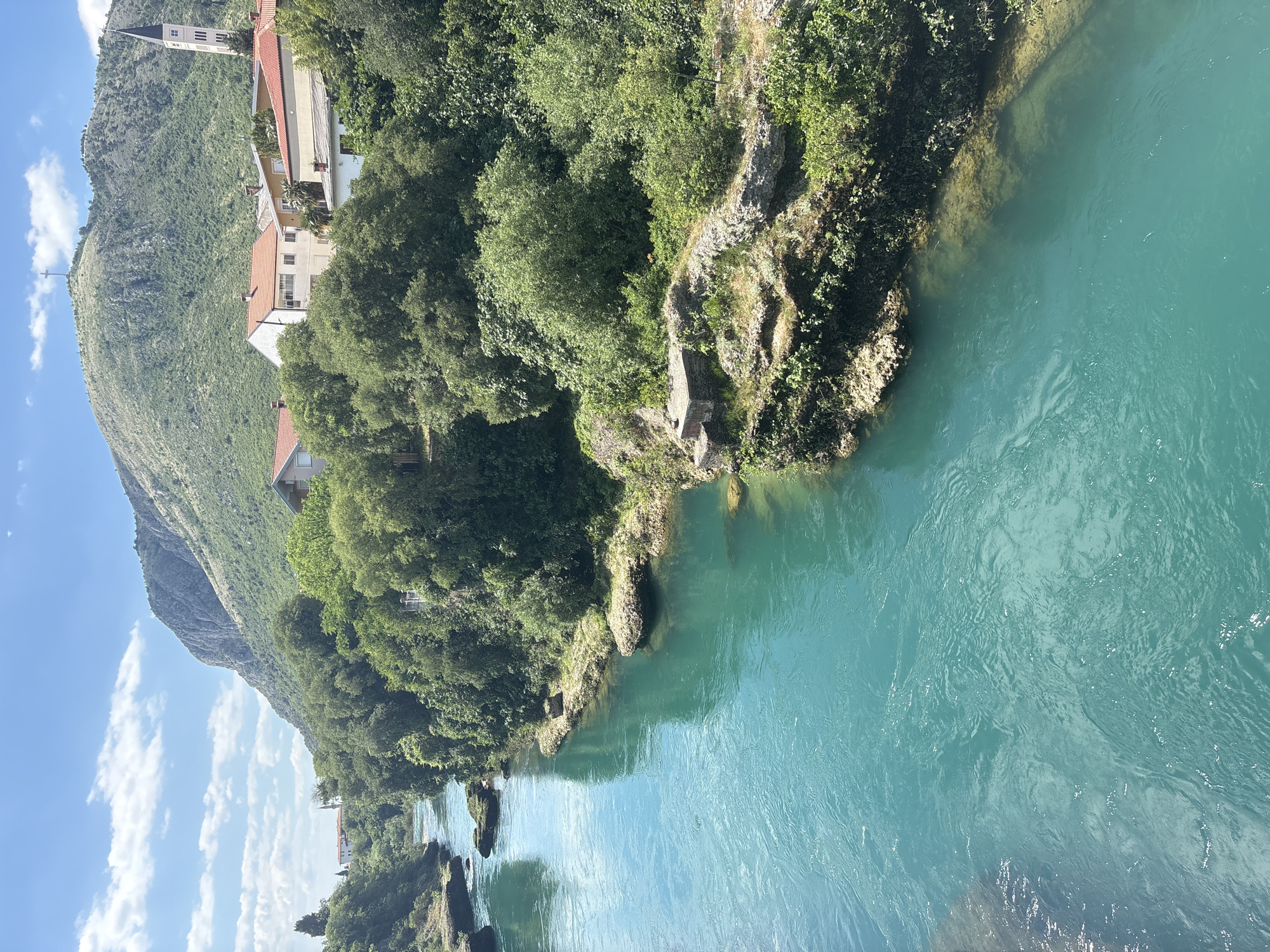 The image captures a segment of the Neretva River in Mostar, Bosnia and Herzegovina, as viewed from an elevated position, possibly a bridge or cliffside. The river's water is a striking turquoise color, suggesting its mineral content and depth. Along the left bank, dense green vegetation dominates, with trees growing along the edge and extending onto a rocky outcropping that juts into the river. Buildings, including some residential structures with red-tiled roofs, are visible amongst the trees further up the slope. In the background, a prominent mountain rises, also covered in vegetation, with a tall, slender structure, possibly a church or tower, visible at its base. The sky is light blue with scattered white clouds, indicating a clear or partly cloudy day. The composition is vertically oriented, providing a sense of depth and emphasizing the river as the central element of the scene.