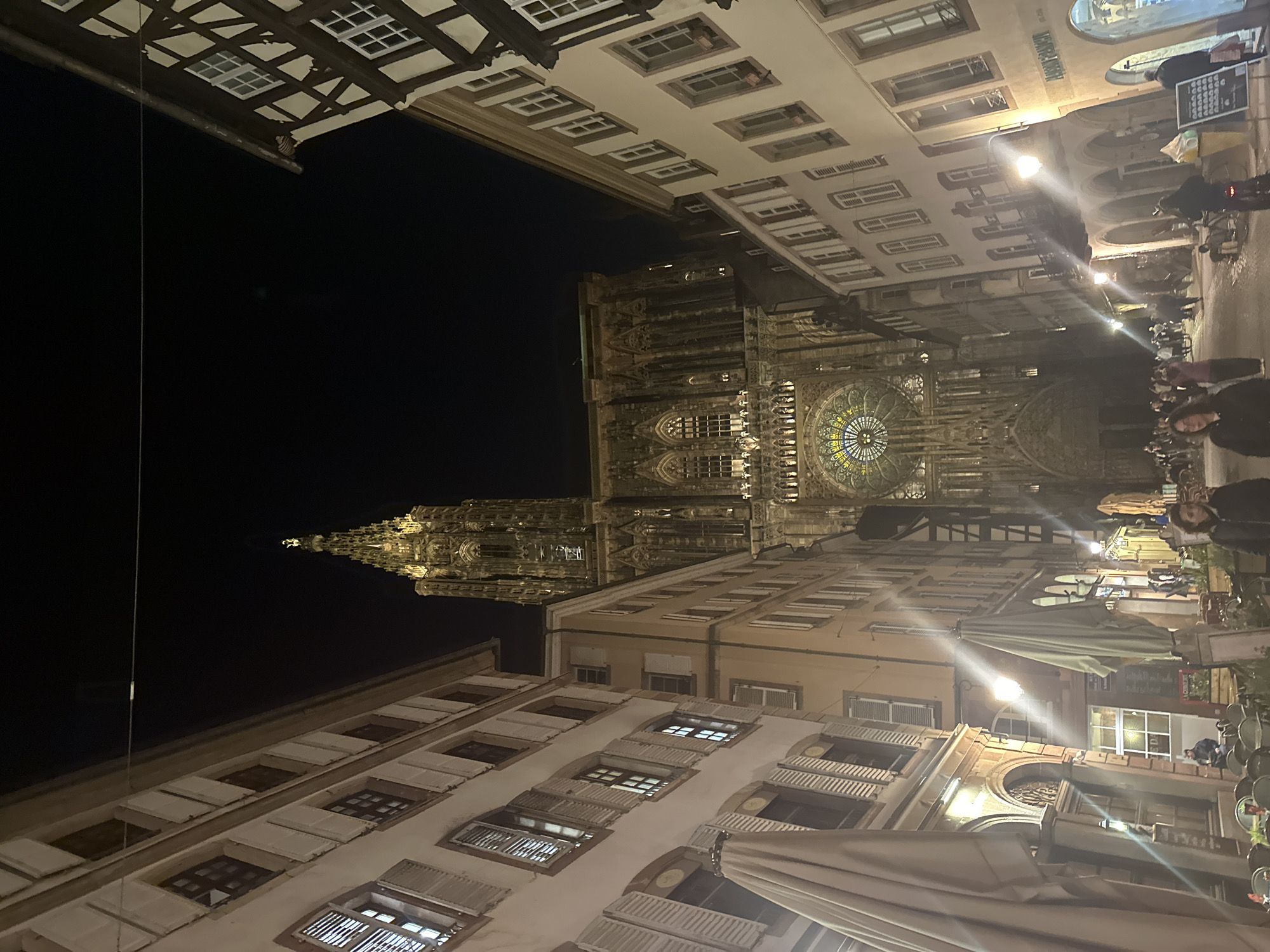 The image is a night shot of a street scene in Strasbourg, France, dominated by the Cathédrale Notre-Dame de Strasbourg in the background. The cathedral is illuminated, highlighting its intricate architectural details, including its spire and rose window. The street is lined with buildings with shutters and windows. People are visible on the right side of the image, suggesting a lively atmosphere. A streetlamp emits a strong light, causing glare. The composition includes the tops of umbrellas, indicating outdoor seating areas for cafes or restaurants.