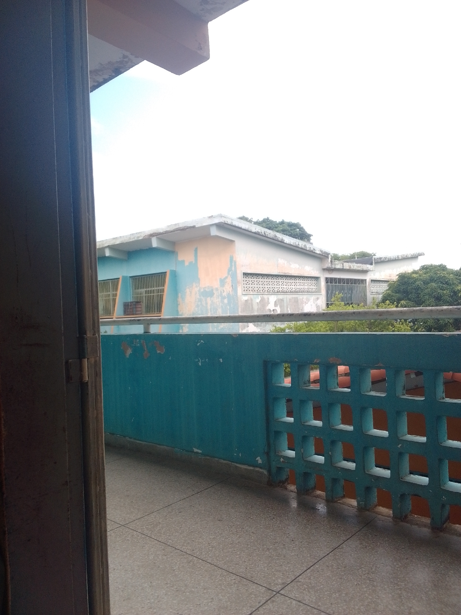 The image depicts a view from an open doorway, looking out onto a weathered building and a tiled patio. The building in the background is painted in pastel blues and oranges, with sections of peeling paint revealing the concrete underneath. It has multiple windows, some with metal bars and others with decorative concrete latticework. Lush green trees are visible behind and around the building, suggesting a tropical or subtropical climate.
In the foreground, a teal-colored, concrete block wall with square openings runs along the edge of the patio. The patio itself is tiled with a speckled pattern and shows signs of recent moisture, possibly from rain. To the left, the dark-colored frame of the doorway is visible, with its hinges suggesting a closed door. The sky appears overcast and bright, indicating daytime, but the diffused light suggests cloudy weather. The overall scene evokes a sense of an ordinary day in a residential or institutional setting. There is no visible text in the image. The location context of Santa Ana, Venezuela, implies a warm climate.