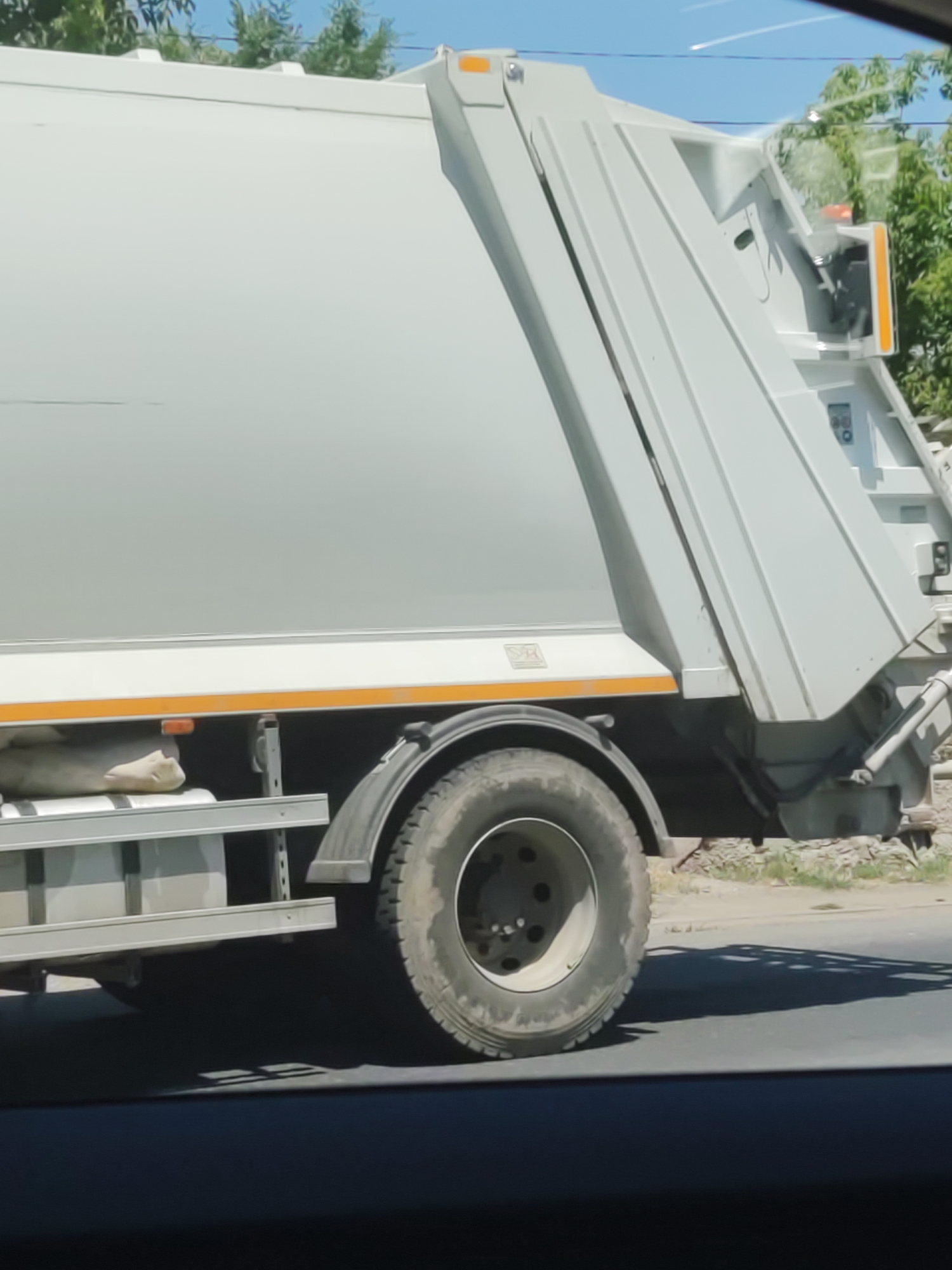 A side view of a white garbage truck is captured, predominantly showing its large, gray container and rear wheel. The truck is on a paved road, with a grassy area and some trees visible in the background. The sky is clear and blue with some faint white lines that could be contrails. The truck appears to be stationary or moving slowly. There are no people visible in the image. The scene is a typical urban or suburban environment, possibly on the outskirts of a city. Given the request to include the location of Shymkent, Kazakhstan, it is plausible this image was taken in that city.