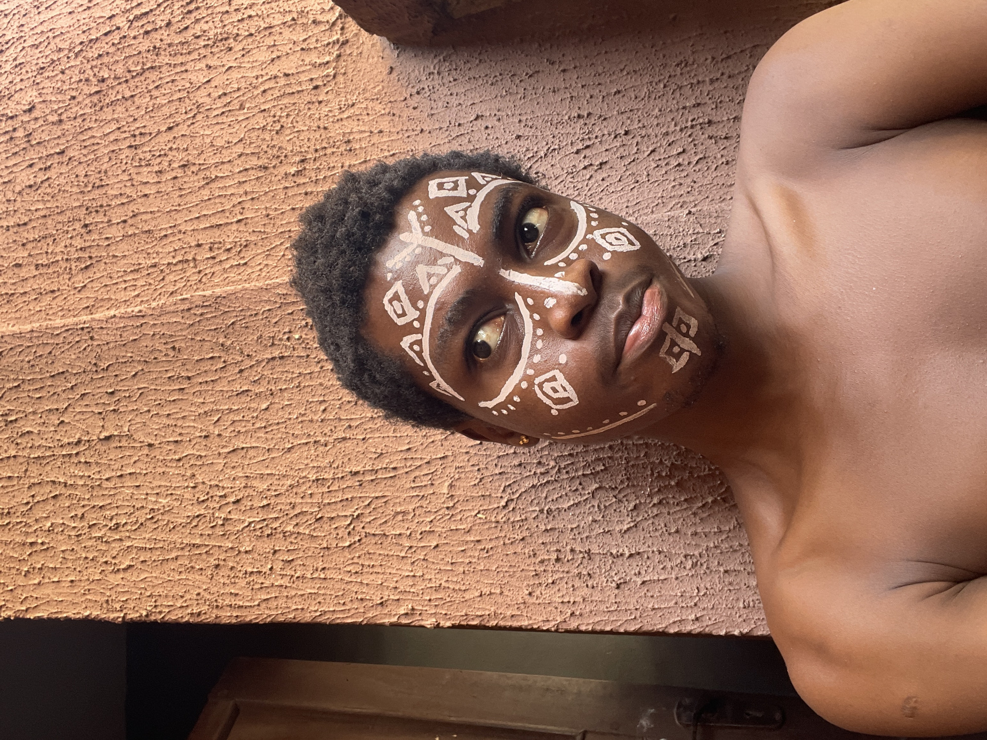 A shirtless dark-skinned man with short curly hair and white geometric face paint is depicted in a close-up, slightly angled portrait. He is lying on a surface with a textured brown wall behind him. A gold earring is visible in his left ear. The bottom of the frame shows a dark wood surface. The scene is located in Nkpor, Nigeria.