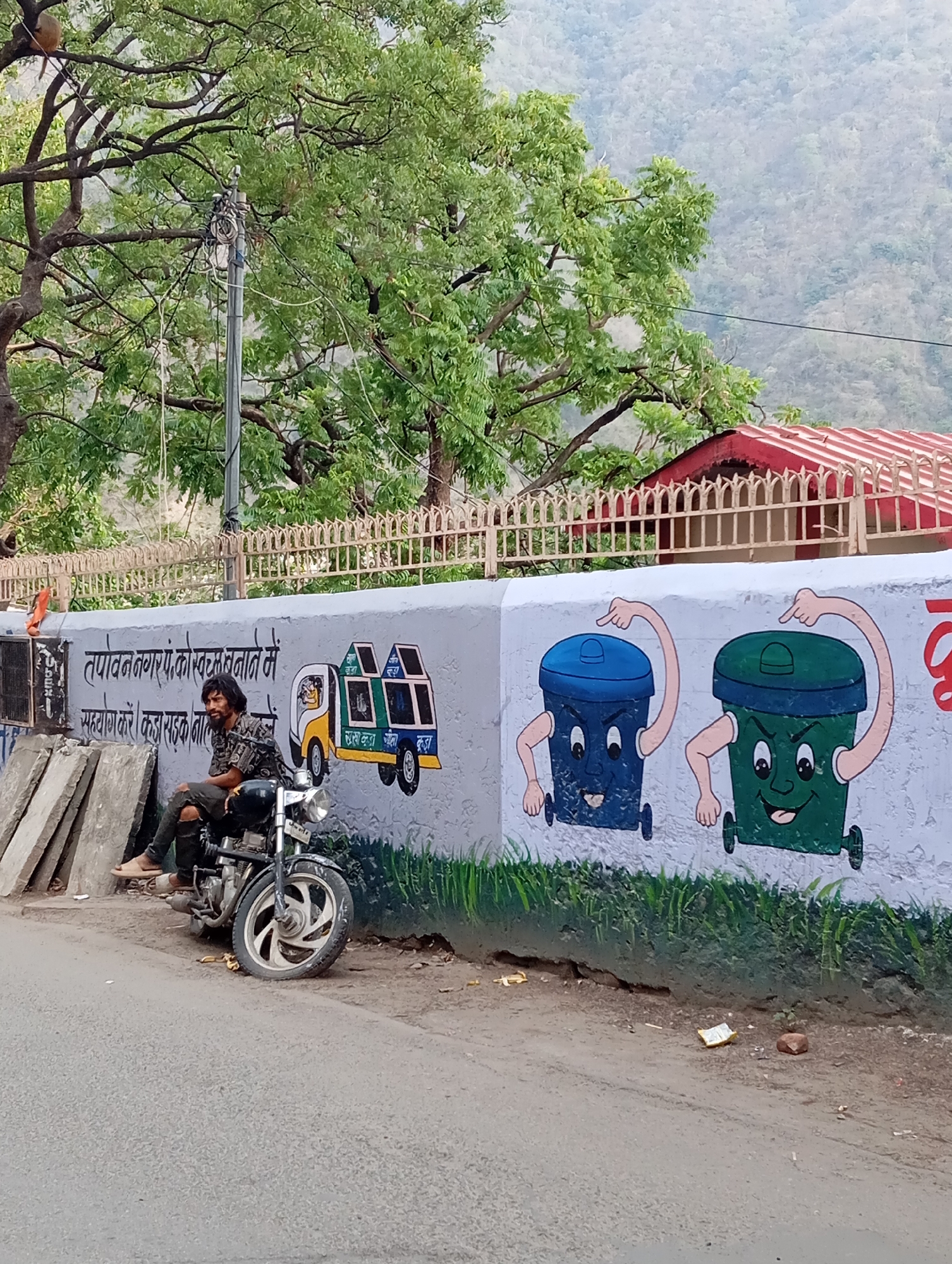The image shows a man sitting on a motorcycle parked next to a painted wall along a street in Unknown, Unknown. The wall features cartoon-like depictions of a garbage truck and two anthropomorphic trash bins. The man has long dark hair and is wearing a patterned shirt, ripped jeans, and sandals. The motorcycle is black with chrome accents. The street is paved, and there is vegetation and a building with a red roof in the background. Overhead power lines and a utility pole are visible, and the landscape includes distant hills. The general atmosphere is daytime and possibly rural.