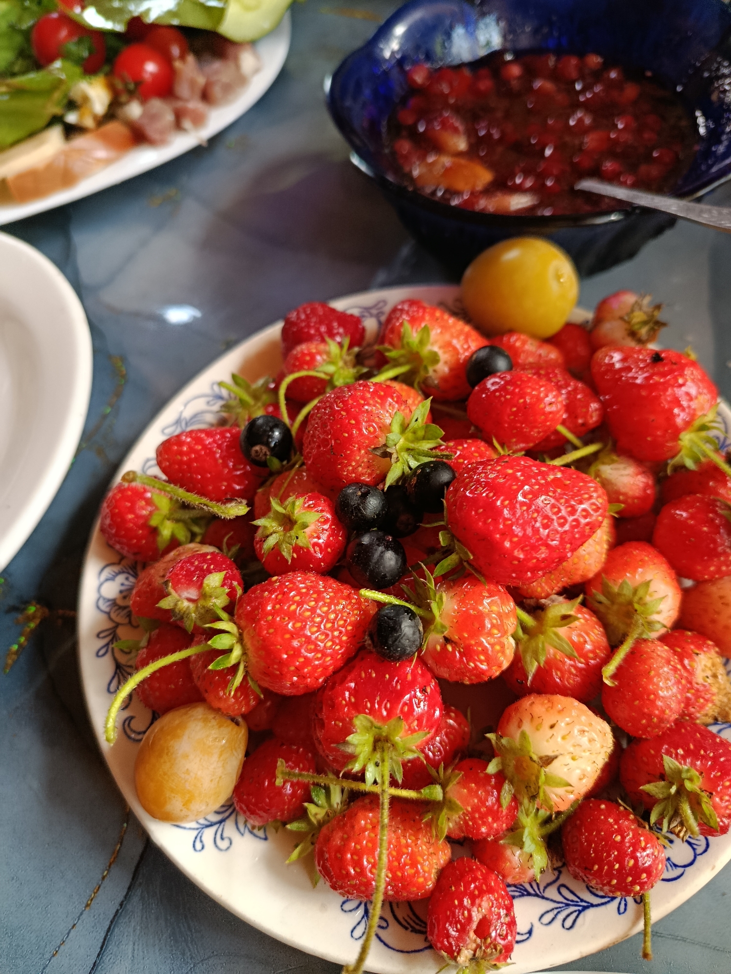 A close-up shot shows a plate overflowing with ripe strawberries and a few dark berries, possibly blueberries or currants. The strawberries are a vibrant red, with some lighter, unripe or pale ones mixed in. The berries are scattered across a white plate with a blue, intricate floral pattern around the rim. A single yellow plum or small fruit sits amongst the berries. In the background, out of focus, a blue bowl filled with a dark, red jam or compote is visible, with a silver spoon resting inside. To the left, a portion of another plate with food, including cherry tomatoes and sliced meat, is slightly visible. The scene appears to be set on a textured, blue surface, possibly a wooden table. The lighting is natural and suggests an indoor setting, likely a home or a cafe. The location is not specified in the image itself.