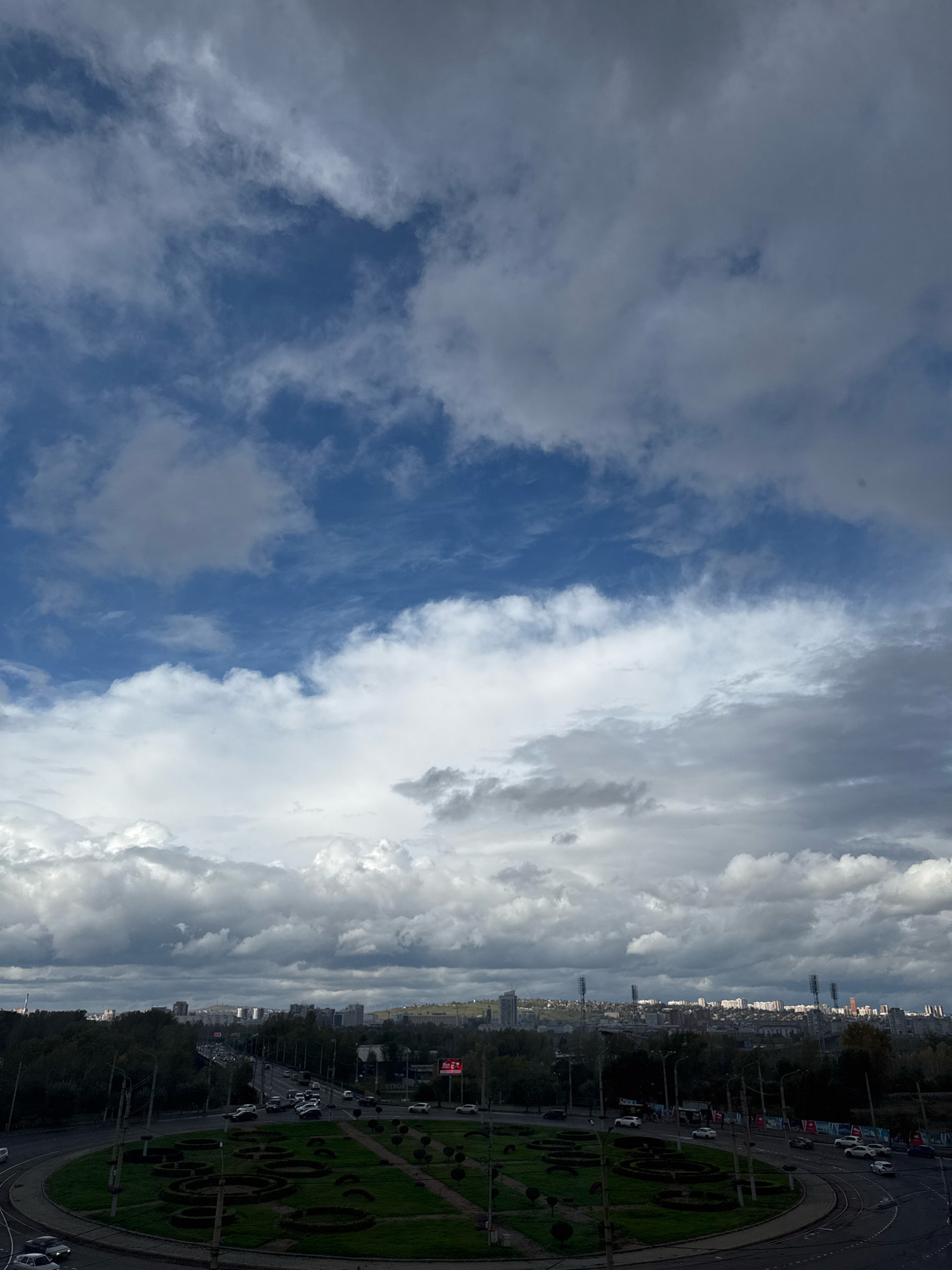 The image depicts a view of a city with a roundabout in the foreground. The roundabout is surrounded by greenery and is traversed by several cars. In the background, the city skyline is visible, with buildings and trees. A bridge with traffic can be seen in the middle ground. The sky is overcast with clouds, with patches of blue sky visible. This scene is located in Krasnoyarsk, Russia, specifically the area around the Karl Marx Square or the adjacent ring road, characterized by the distinctive roundabout and the surrounding urban landscape. No people are clearly visible in the image.