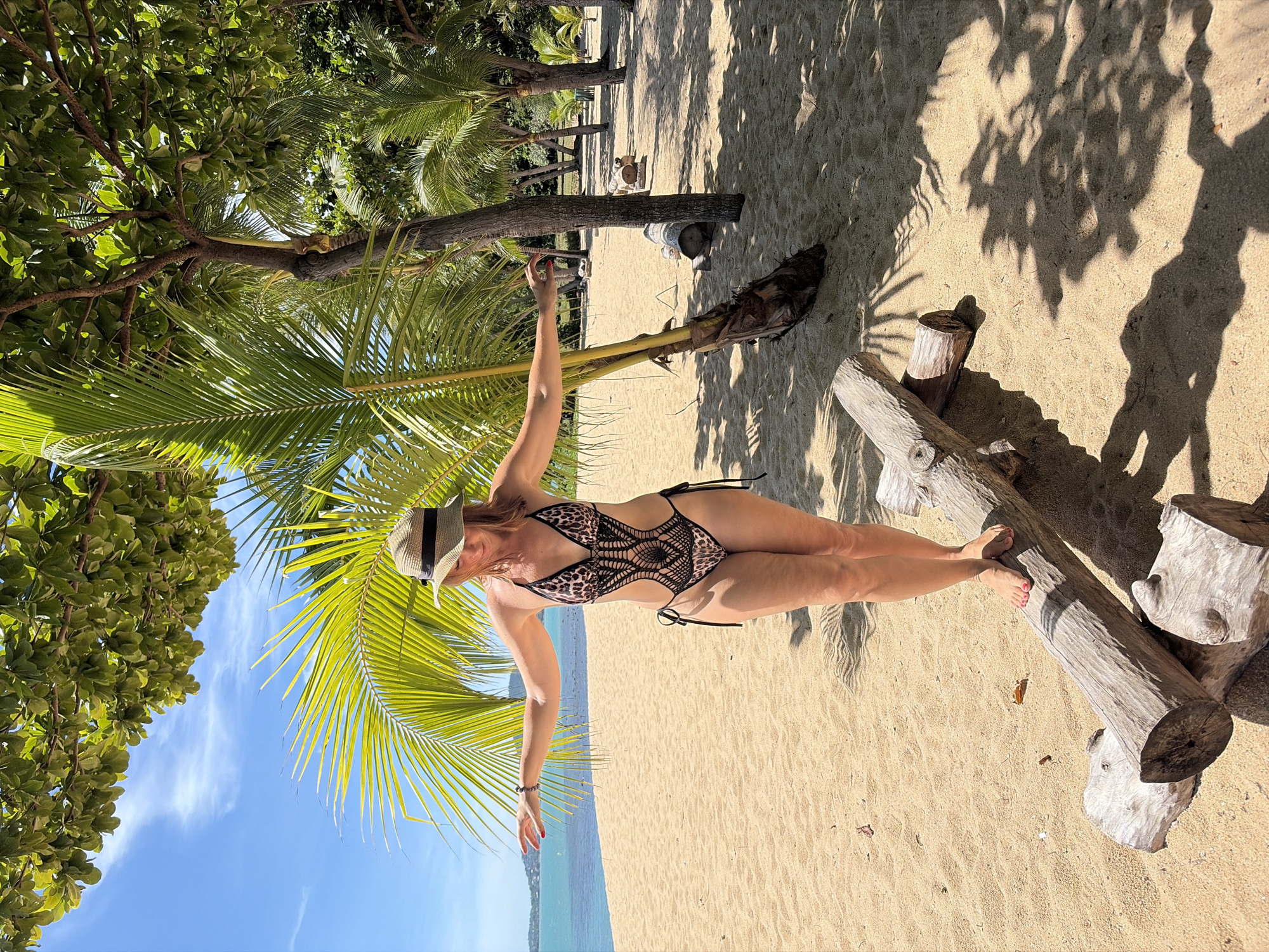 A woman wearing a leopard-print one-piece swimsuit and a straw hat stands on a log on a sandy beach. She has her arms outstretched to her sides, balancing herself. She is looking down and to her left. Behind her, palm trees line the beach, and the ocean is visible in the distance. The sky is blue with some white clouds. The image was taken in Baan Bo Phut, Thailand.
