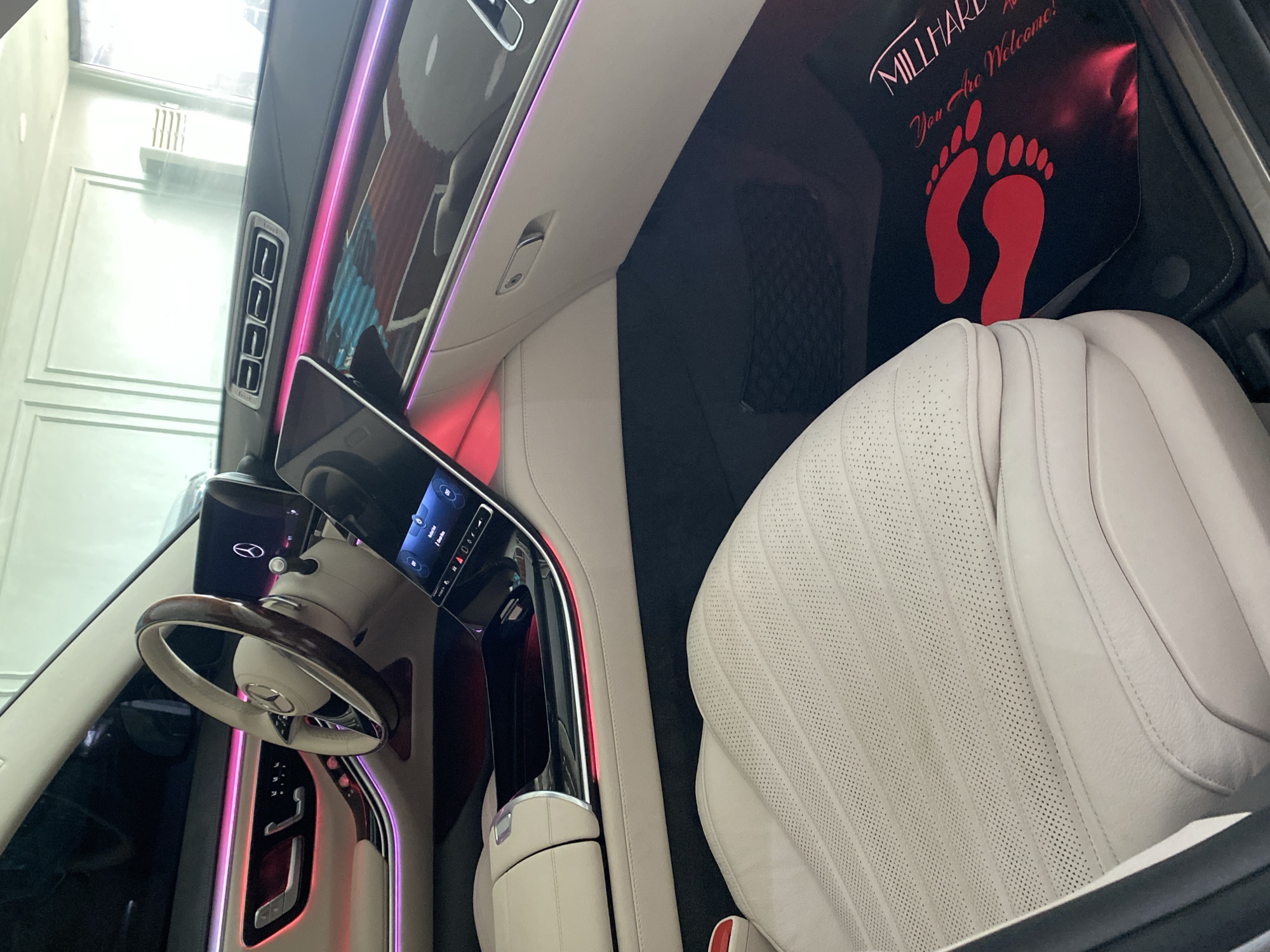 A partial view of the interior of a luxury car, likely a Mercedes-Benz, possibly located in Igboefon, Nigeria (based on the "MILLHARD" floor mat which could be a local service). The scene is brightly lit, showing a white leather seat with perforated detailing, a dashboard with a digital display and ambient lighting (pink and red), and a steering wheel with wood trim. A black floor mat with red footprints and the text "MILLHARD You Are Welcome!" is visible. The car appears to be in a showroom or well-lit garage. No people are visible.