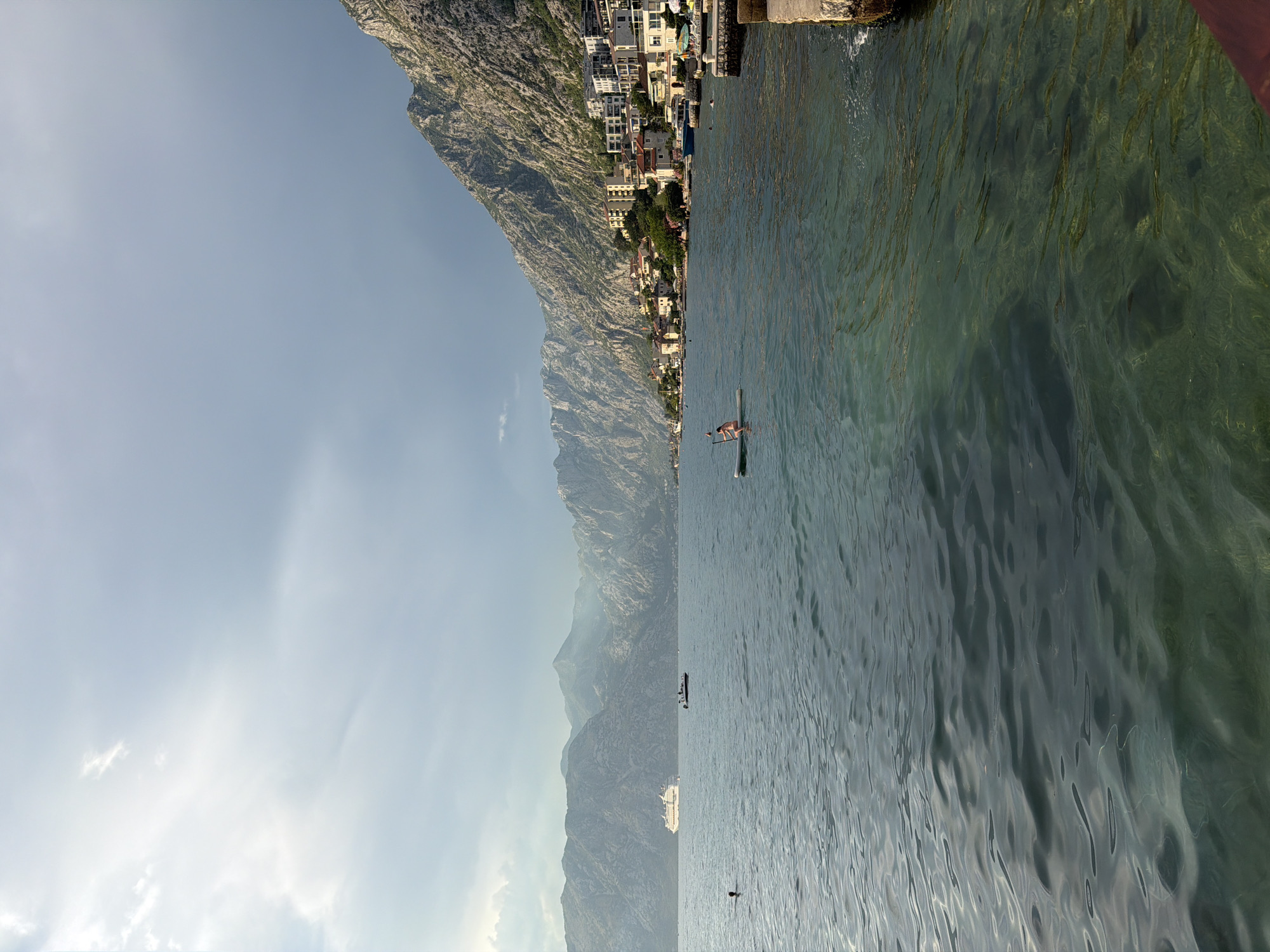 The image depicts a scenic view of the Bay of Kotor in Dobrota, Montenegro, characterized by a calm, turquoise sea meeting steep, rocky mountains that are dotted with buildings. The sky above is a blend of light blue and grey clouds, with some sunlight breaking through.
In the foreground, the clear water reveals the seabed below. A person is seen paddleboarding on the water, facing the shore. Further out in the bay, a large cruise ship is visible on the left, and a smaller motorboat with passengers is positioned between the cruise ship and the shore.
The middle ground features the coastline of Dobrota, with a collection of residential buildings and hotels nestled against the mountainside. A promenade or walkway lines the edge of the water, with some stone structures and possibly seating areas.
The background is dominated by the imposing, rugged mountains that form the backdrop of the bay. The sheer scale of the mountains and their textured surfaces are evident. The overall atmosphere is serene and picturesque, highlighting the natural beauty of the location combined with human habitation.