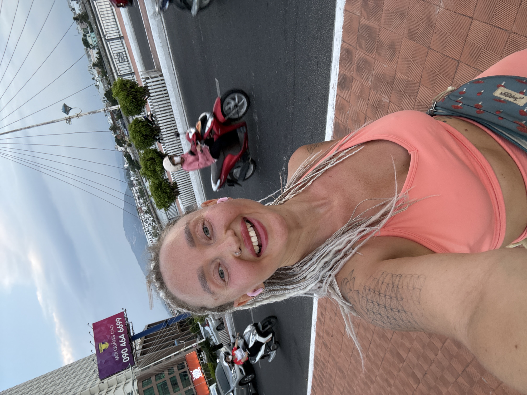In Nha Trang Ward, Vietnam, a woman with blonde braided hair and pink earbuds takes a selfie while smiling. She is wearing a coral-colored sports bra and matching shorts. A fanny pack with a mushroom pattern is visible around her waist. In the background, a red scooter carries a person wearing a helmet and a pink shirt. Other scooters and cars are on the road, and a tall building with a billboard displaying "MỌI QUẢNG CÁO 090 464 6699" is to the left. The scene appears to be near a bridge with a network of cables overhead. The ground is paved with reddish-brown tiles.