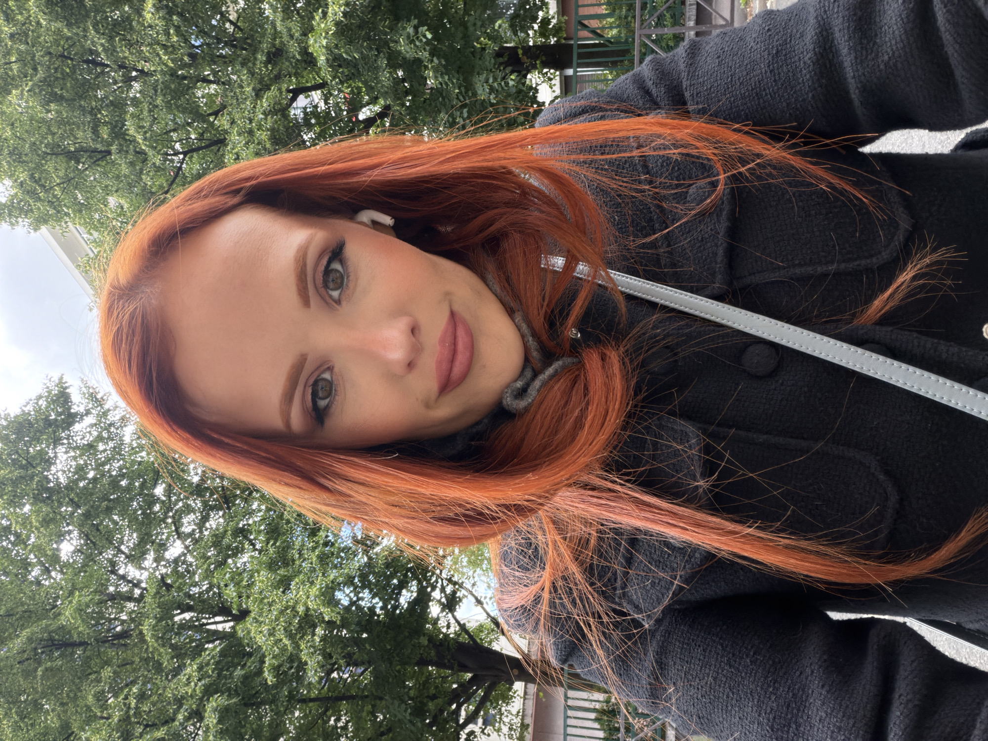 A young woman with reddish-orange hair and fair skin is facing the camera. She is wearing a dark grey wool coat over a grey turtleneck sweater. An earbud is visible in her left ear. She appears to be outdoors, with a tree with green foliage visible in the background. A fence is also visible in the background. The image is taken at an angle. The exact location in Paris, France cannot be determined from the given image.