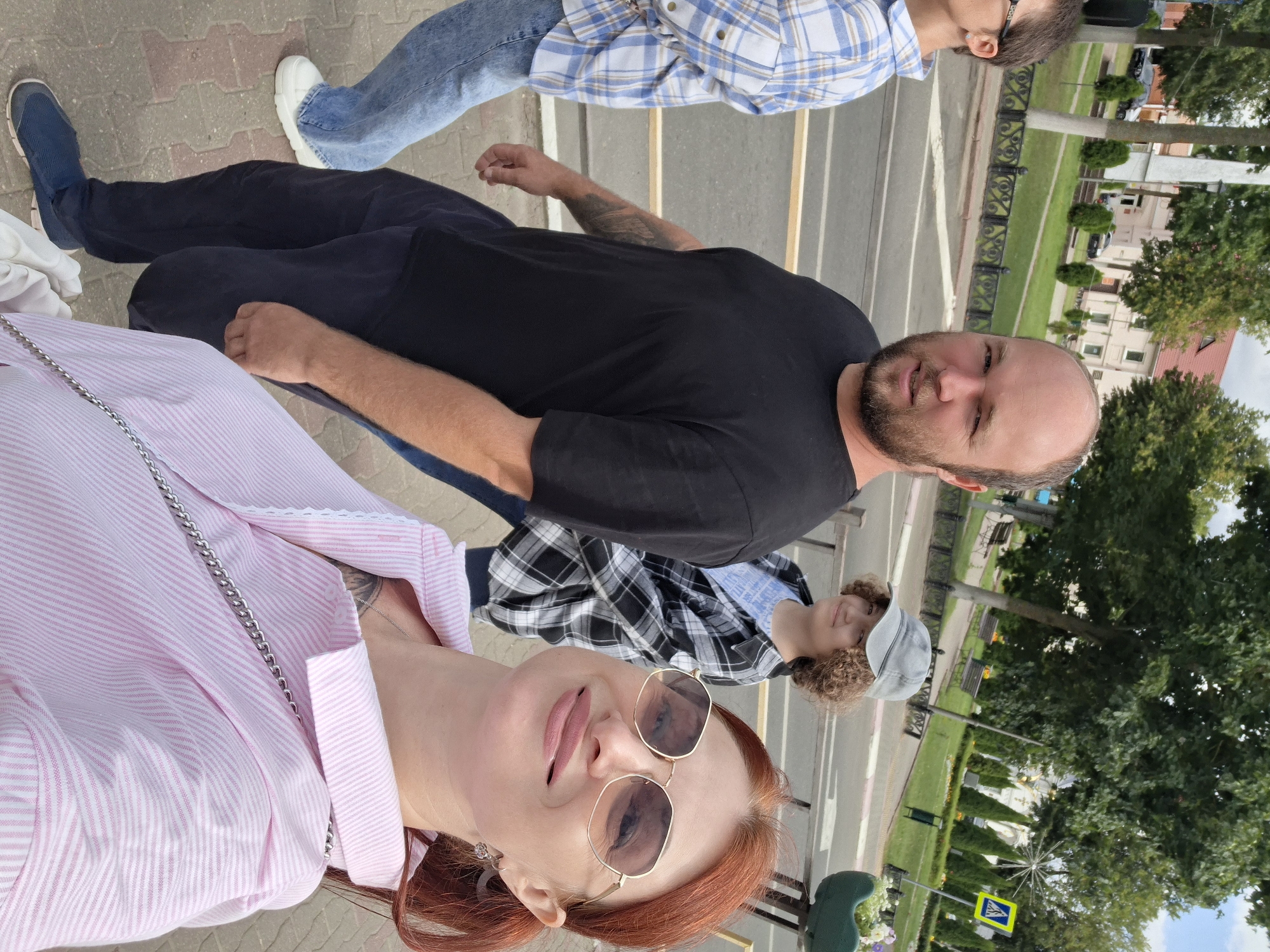 In Pastavy, Belarus, a group of four individuals, two adults and two children, are captured in a candid outdoor photograph, likely during daytime. The composition focuses on a man with a beard and a woman with red hair and sunglasses in the foreground, while two children are visible in the background. The individuals appear to be walking on a sidewalk adjacent to a road, with a park-like area featuring trees, benches, and a building visible across the street. The atmosphere suggests a casual outing in an urban setting.
