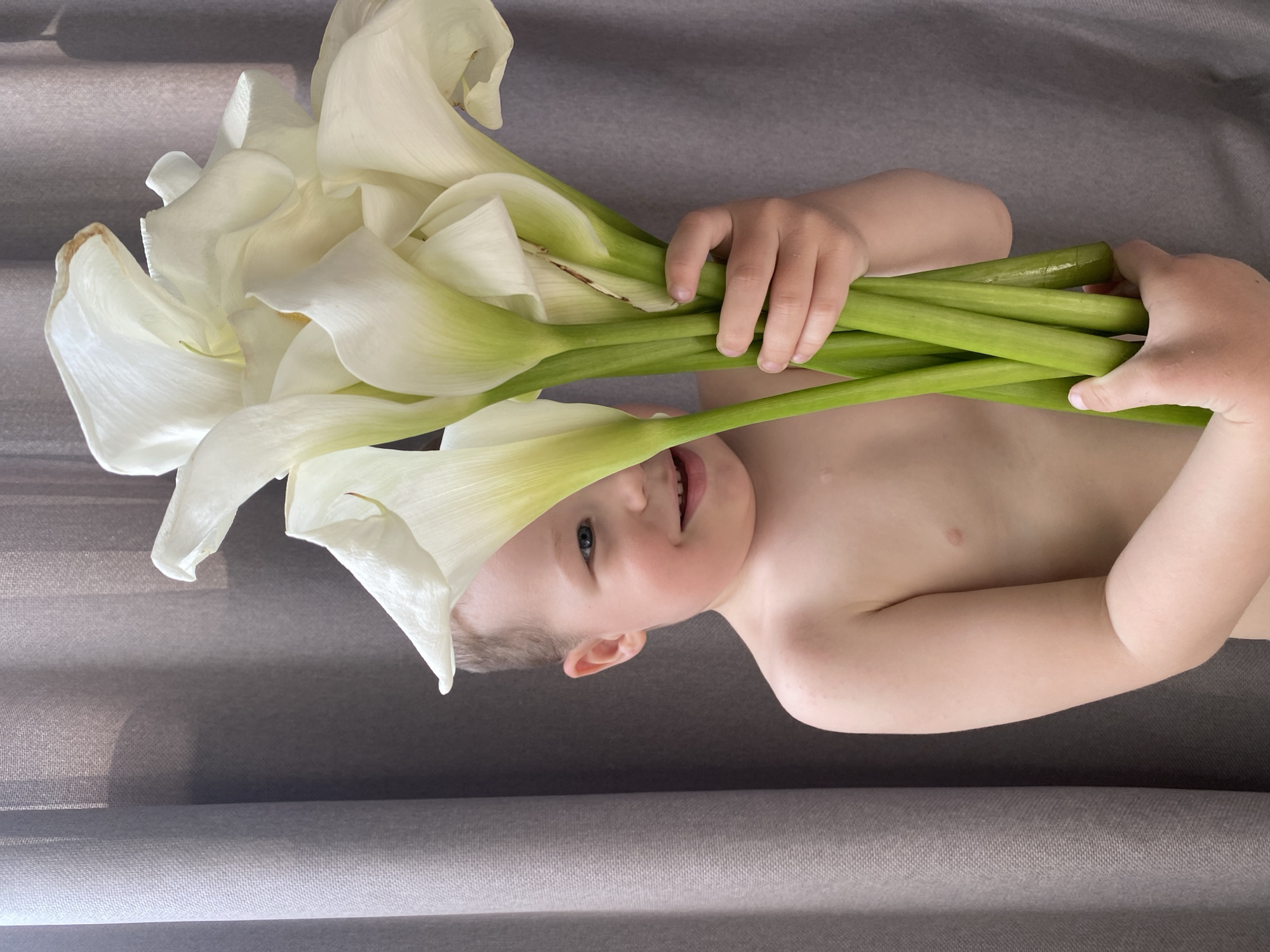 A young boy, possibly Caucasian, is holding a bouquet of white calla lilies in front of a grey curtain. He is partially obscured by the flowers, with only his right eye and cheek visible. He has fair skin, light brown hair, and is smiling. His shoulders and upper chest are bare. The calla lilies have thick green stems and large, white trumpet-shaped flowers. The background is a plain grey fabric, likely a curtain, with some soft shadows suggesting natural light from the right. The image appears to be a candid portrait. There is no indication of the city of Sochi, Russia, or any specific urban environment.