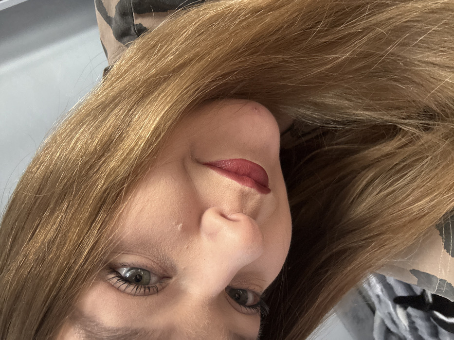 The image depicts a close-up, tilted portrait of a female individual, photographed in an indoor setting in Chaikovsky, Russia. The subject's head is angled approximately 45 degrees counter-clockwise, with her gaze directed towards the viewer. She has light brown to auburn hair, long and straight, which cascades across the right side of the frame. Her eyes are green/hazel, featuring prominent dark eyelashes. Her nose and the lower portion of her face are visible, including her lips which are colored with a dark red/burgundy, matte lipstick and upturned slightly, indicating a faint smile. Her skin appears fair; a small, light-colored blemish is present on her right cheek below the eye, and another on her chin. A portion of a camouflage-patterned garment is visible on her upper right side. The background consists of a light grey surface on the upper left and a textured grey object, possibly a blanket or cushion with a darker pattern, on the lower left.