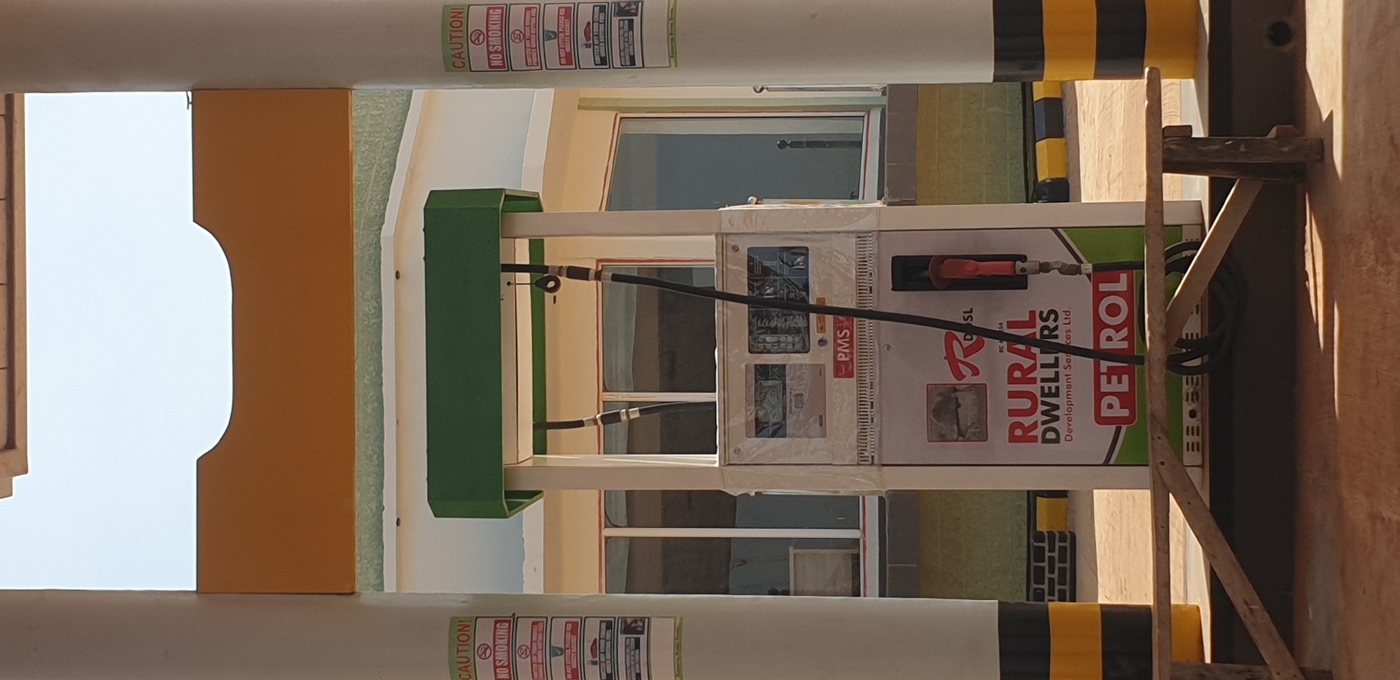 A petrol station pump is shown in the center of the image. It is a multi-purpose pump, with the brand "Rural Dwellers Petrol" visible on the front. The pump is white with green and red accents. There are two hoses attached to the pump, one on the left and one on the right. The pump has a digital display that shows prices and amounts. A red and black nozzle is hanging from the right side of the pump. Two support pillars are visible on either side of the pump. The left pillar has a "CAUTION!" and "NO SMOKING" sign attached to it. The background shows a building with large windows and a light-colored roof. The area around the pump is paved with light brown bricks. A wooden bench is placed in front of the pump. The scene appears to be outdoors on a sunny day. The location is Sabon Tasha, Nigeria, a busy area known for its commercial activities, including a prominent petrol station that serves the local community.