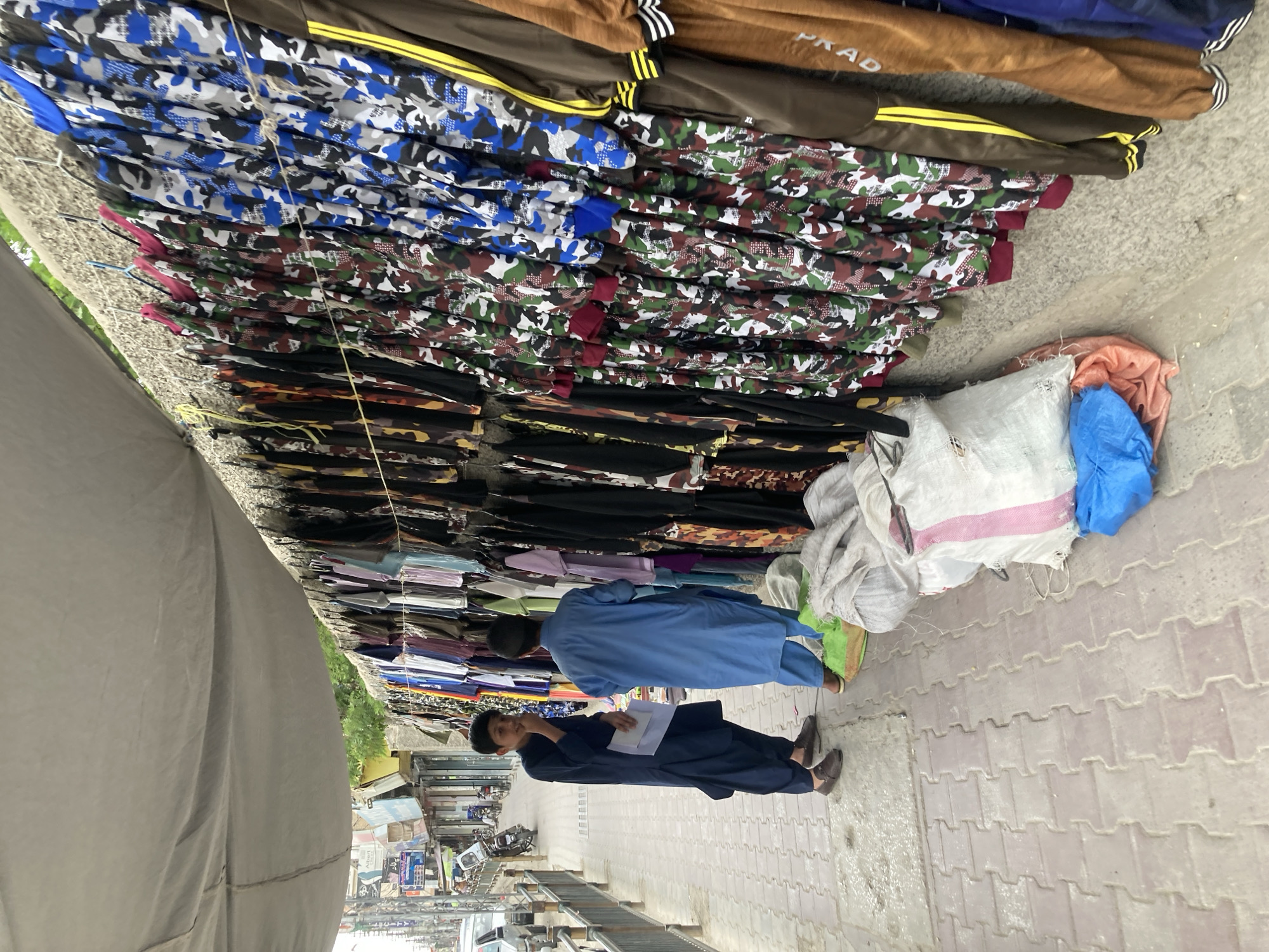 Two men stand on a sidewalk paved with light-colored brick, examining merchandise displayed in a shop or street stall. The man closest to the camera is dressed in a dark blue shalwar kameez and holds white papers in his hands. The other man, further from the camera, wears a lighter blue shalwar kameez and is also looking at the items. The merchandise primarily consists of garments with camouflage patterns, along with some brown and black garments. The garments are hung on hangers and displayed along a wall. To the right of the men, there are some sacks or bags filled with items. The scene shows an open street in Quetta City Tehsil, Pakistan.