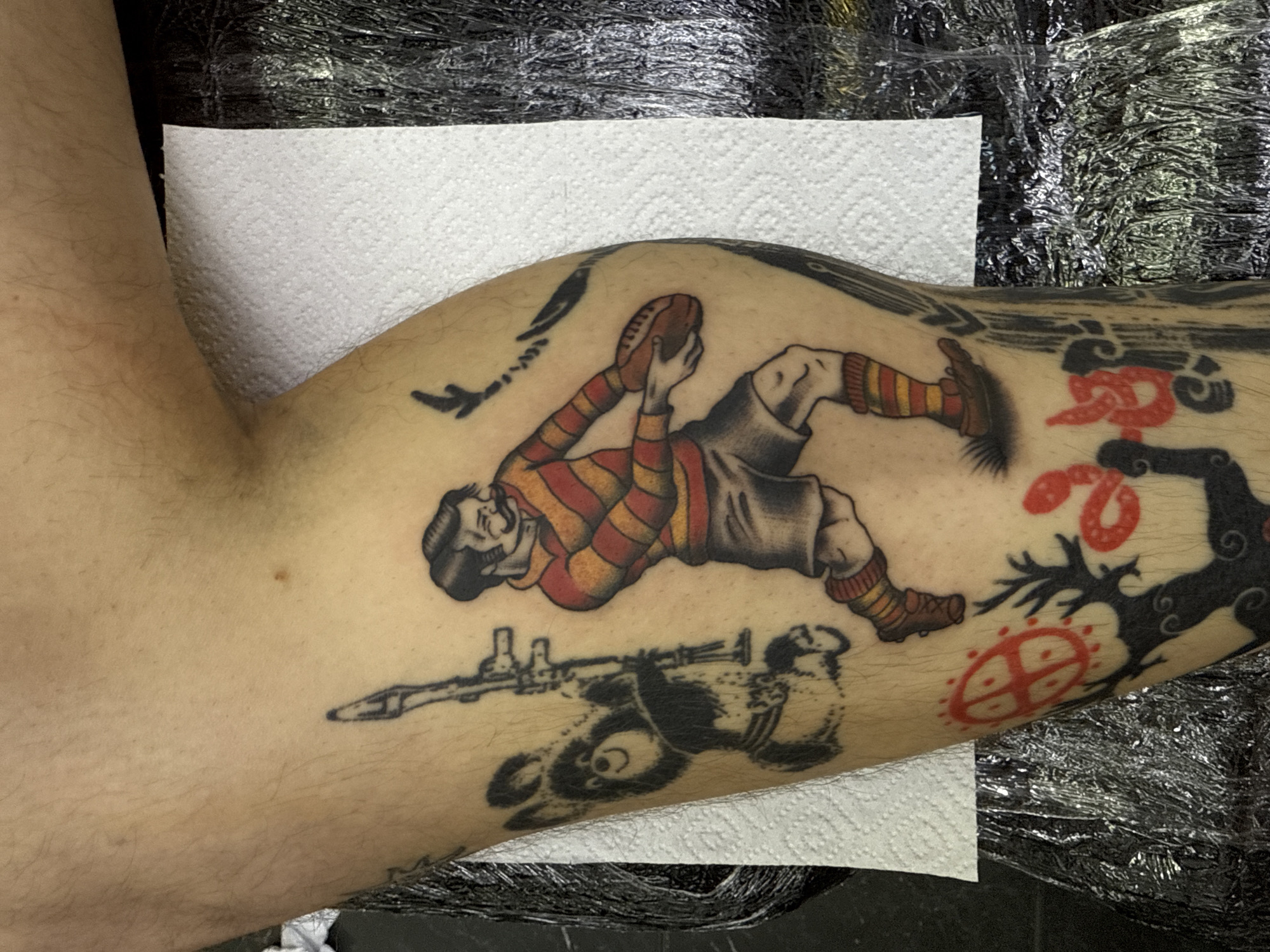 The image captures a close-up view of a person's muscular arm, extensively covered in tattoos, suggesting an indoor setting, possibly a tattoo studio.
The most prominent tattoo features a male rugby player in a dynamic pose, mid-action, holding a brown rugby ball. He wears a traditional striped jersey (red, orange, and yellow), grey shorts, and striped socks with brown shoes. The tattoo style is bold and illustrative, typical of American traditional tattooing, with clear outlines and solid color fills.
Below the rugby player, there is a dark silhouette tattoo of a stylized figure with a prominent, round head and large eyes, appearing to hold a rifle across its body. To the right of the rugby player, an intricate black tattoo of gnarled, tree-like branches or abstract organic shapes extends upwards, interwoven with several vibrant red snake-like or abstract designs. Further down the arm, below the tree-like design, is a circular red emblem featuring a cross pattern and dots within its perimeter. Higher up on the arm, above the rugby player, a small black outline of an airplane is visible.
The skin around the tattoos is light-toned and exhibits fine arm hair. In the background, parts of a white paper towel and crinkled black plastic wrap are visible, suggesting either a fresh tattoo being protected or supplies from a recent tattooing session. No text is visible in the image. The overall impression is a vibrant and diverse collection of traditional tattoo artwork.